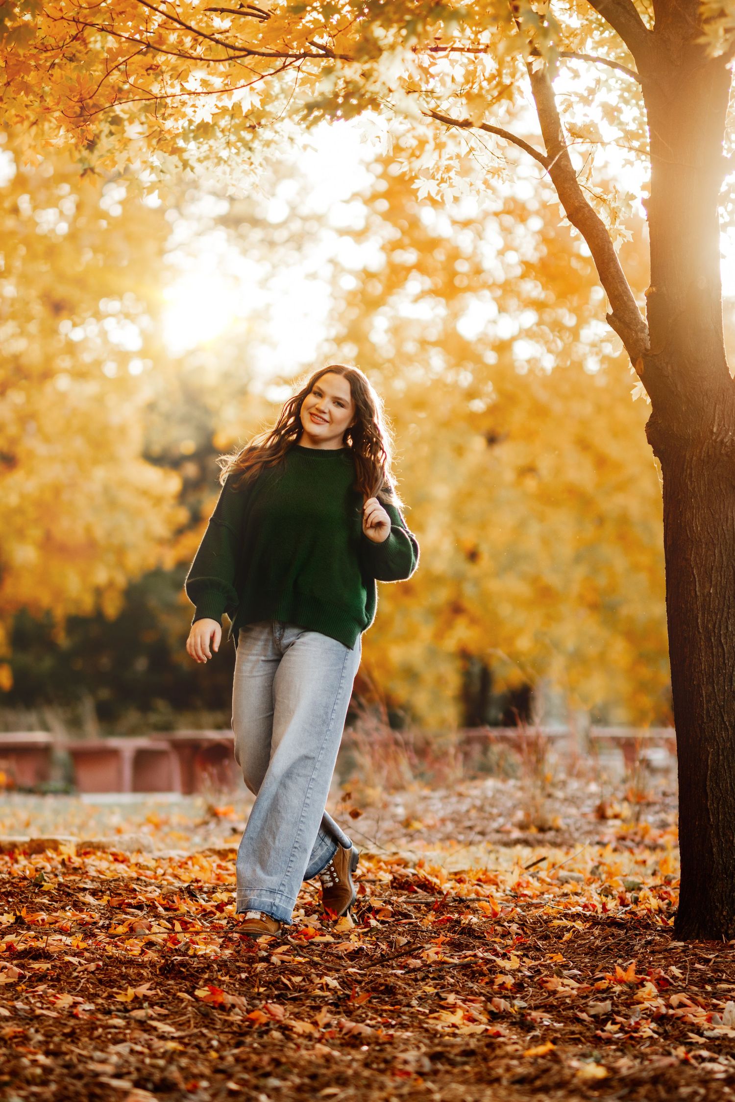 Person in black sweater and jeans playing in golden autumn leaves during sunset in a park setting.