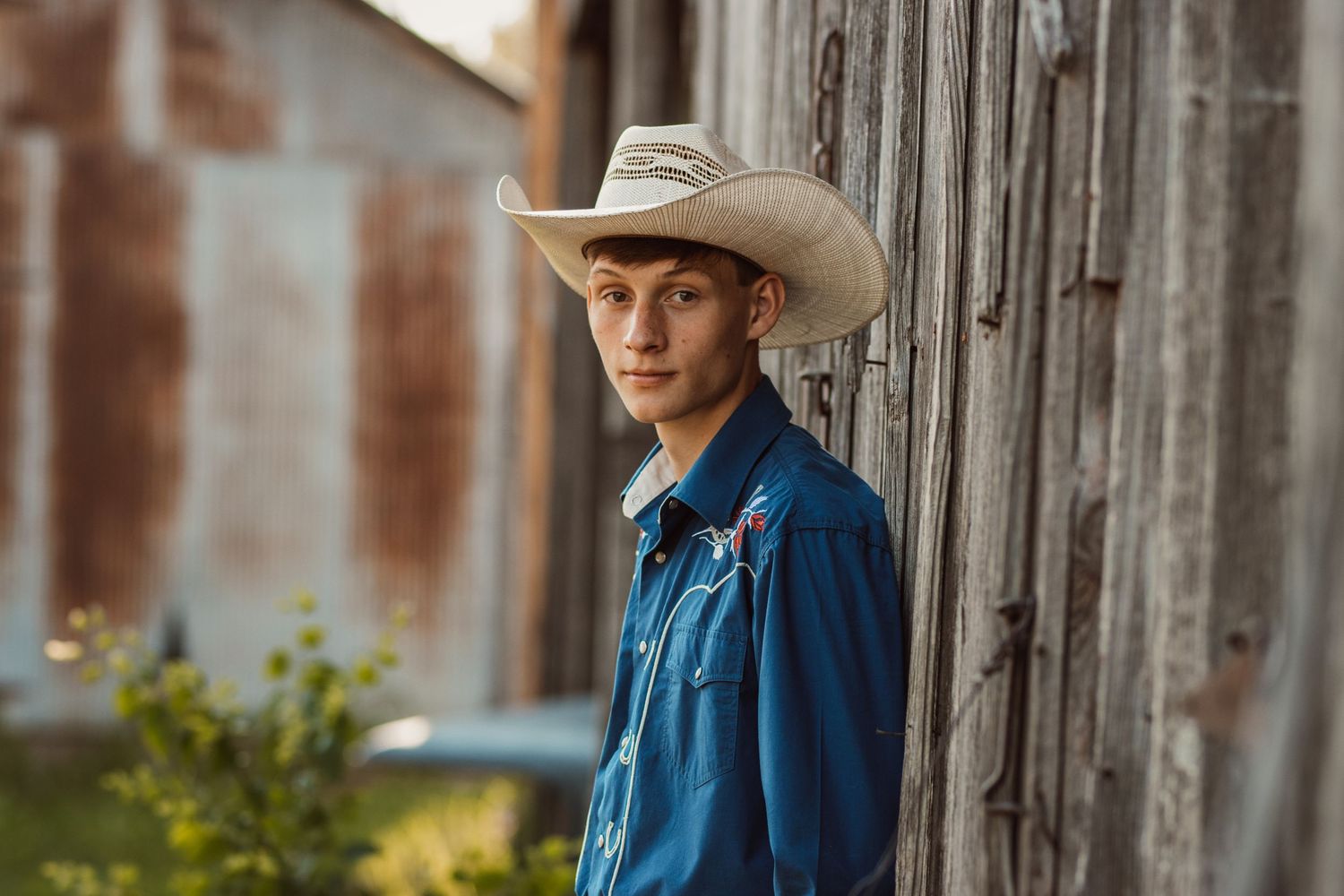 Person in denim shirt and cowboy hat stands against weathered wooden wall.