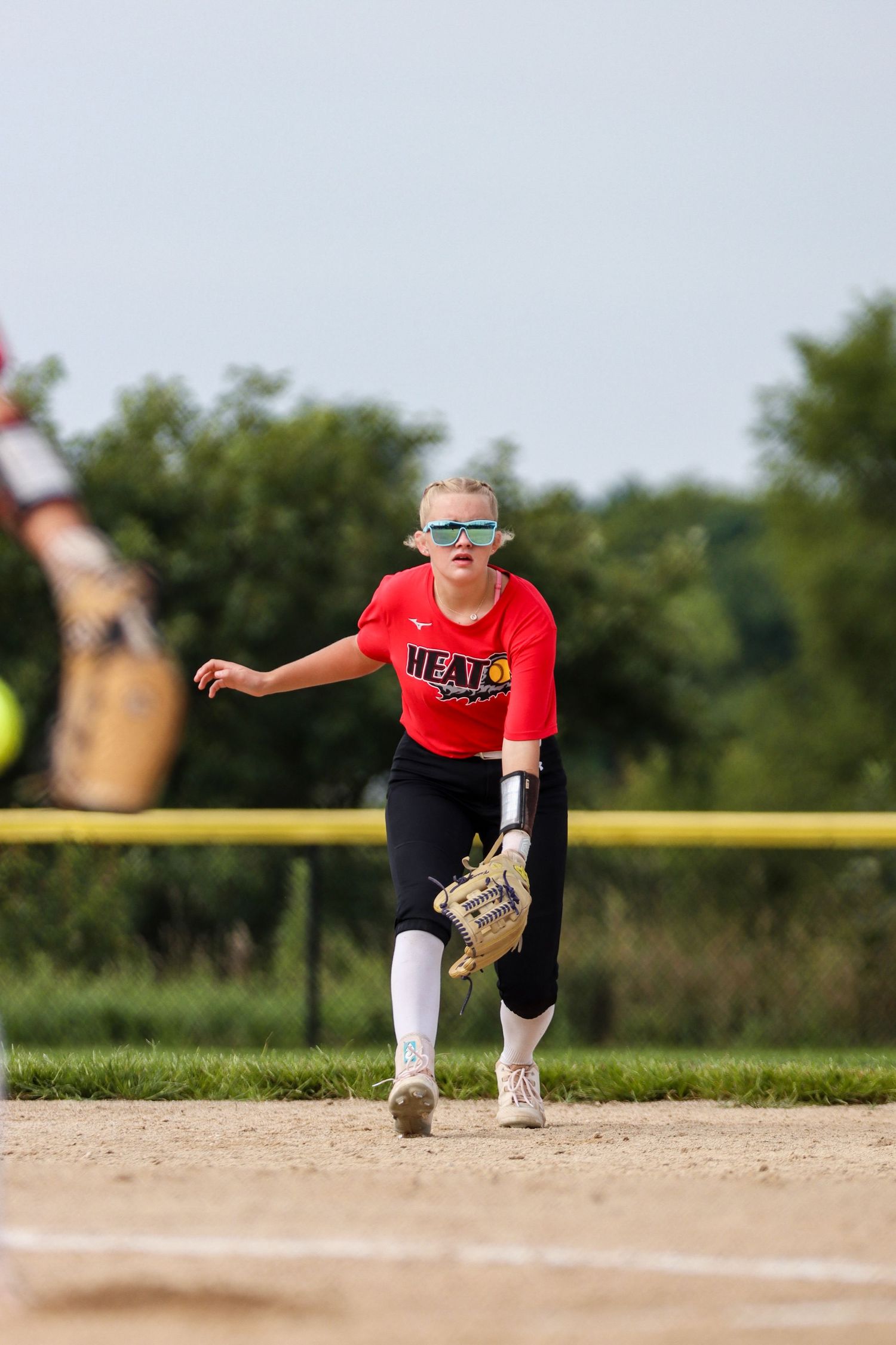 Softball player wearing red jersey and sunglasses winds up for pitch on dirt field.