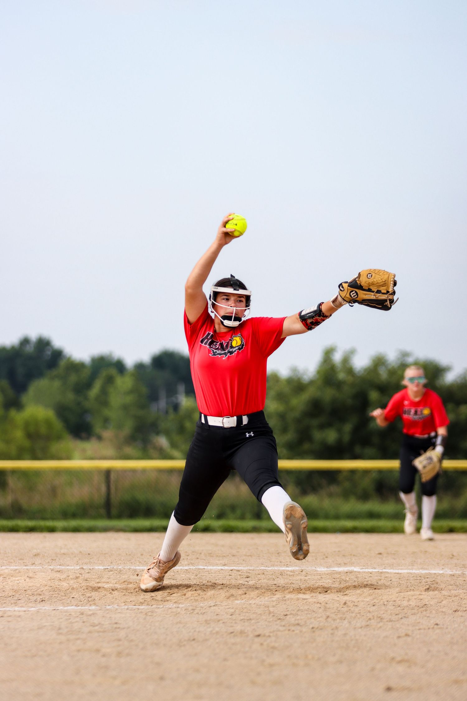 Softball pitcher in red jersey throwing yellow ball during outdoor game on dirt field.