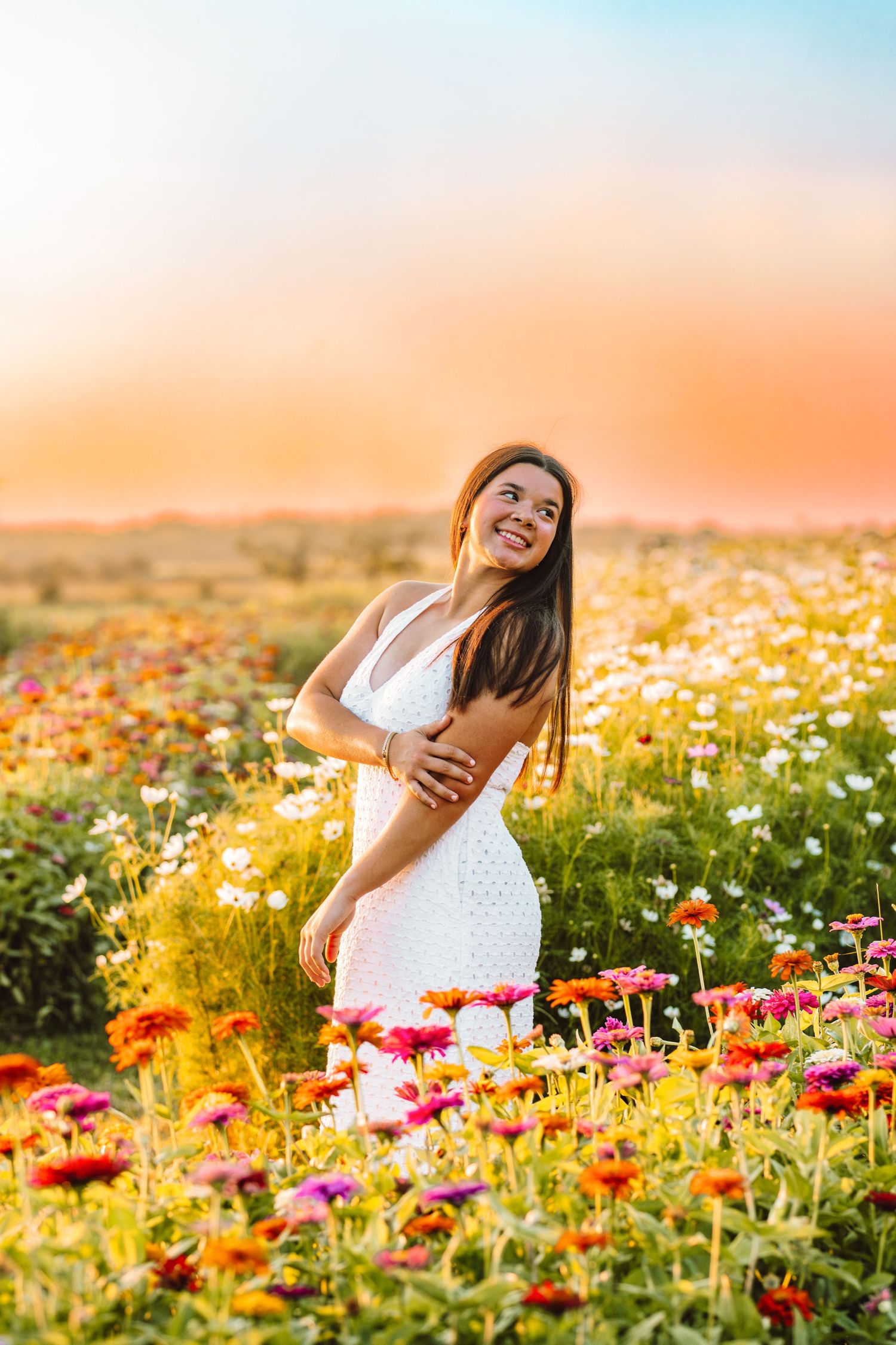A woman in a white dress stands joyfully among vibrant wildflowers during a golden sunset.