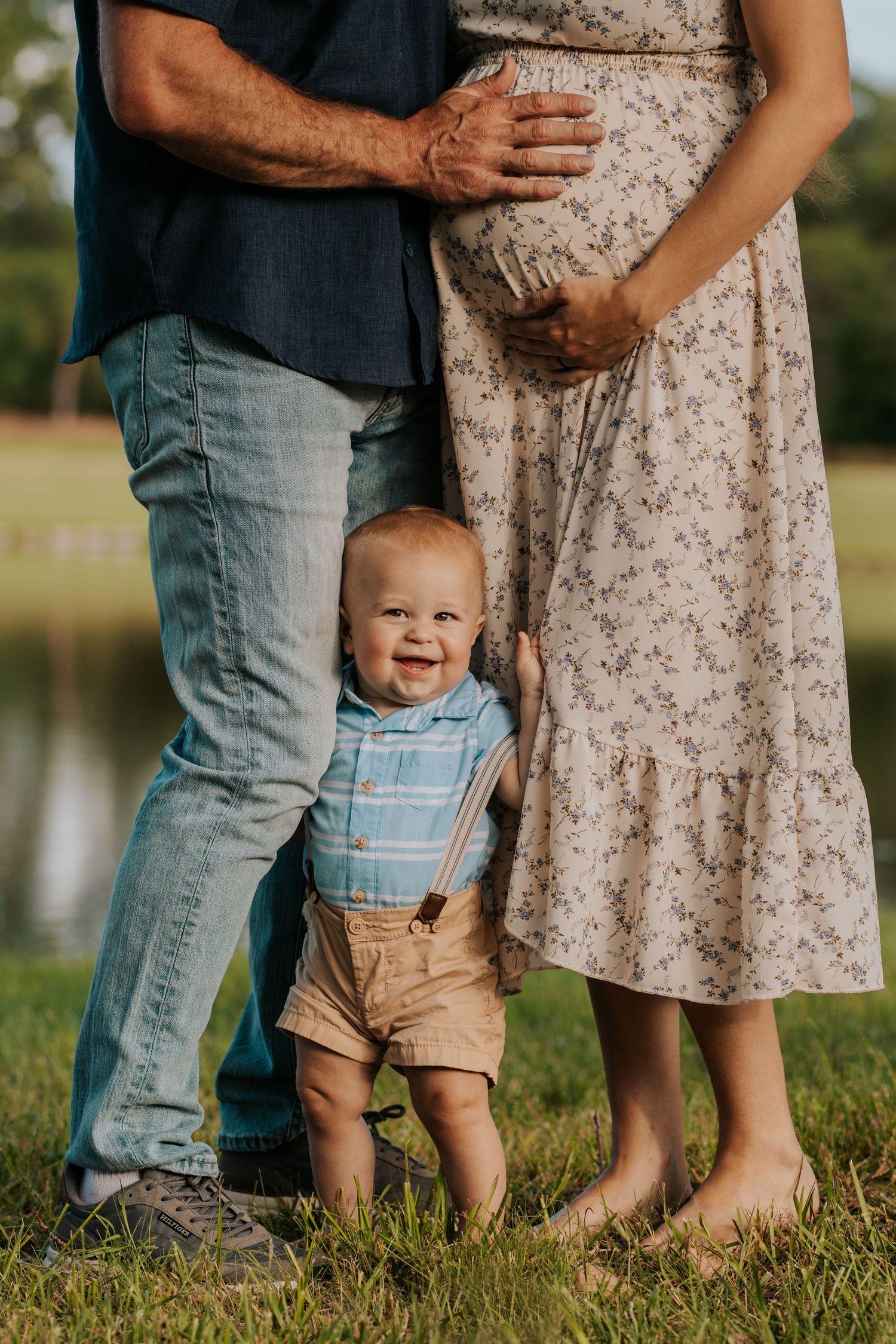 Family maternity photo shows toddler in suspenders standing between parents on grassy lawn near water at sunset.