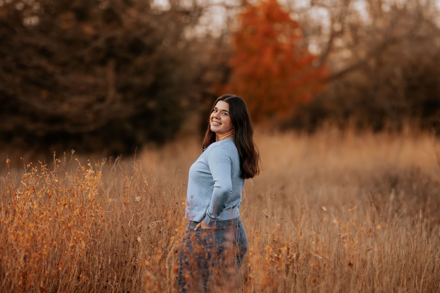 Person in light blue sweater stands in golden autumn field during sunset creating a dreamy outdoor portrait.