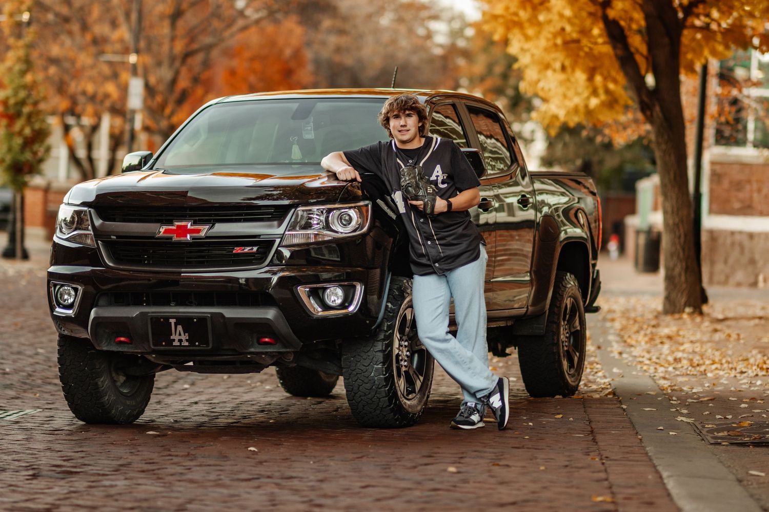 A black Chevrolet Colorado pickup truck parked on a brick path during autumn with fallen leaves on the ground.