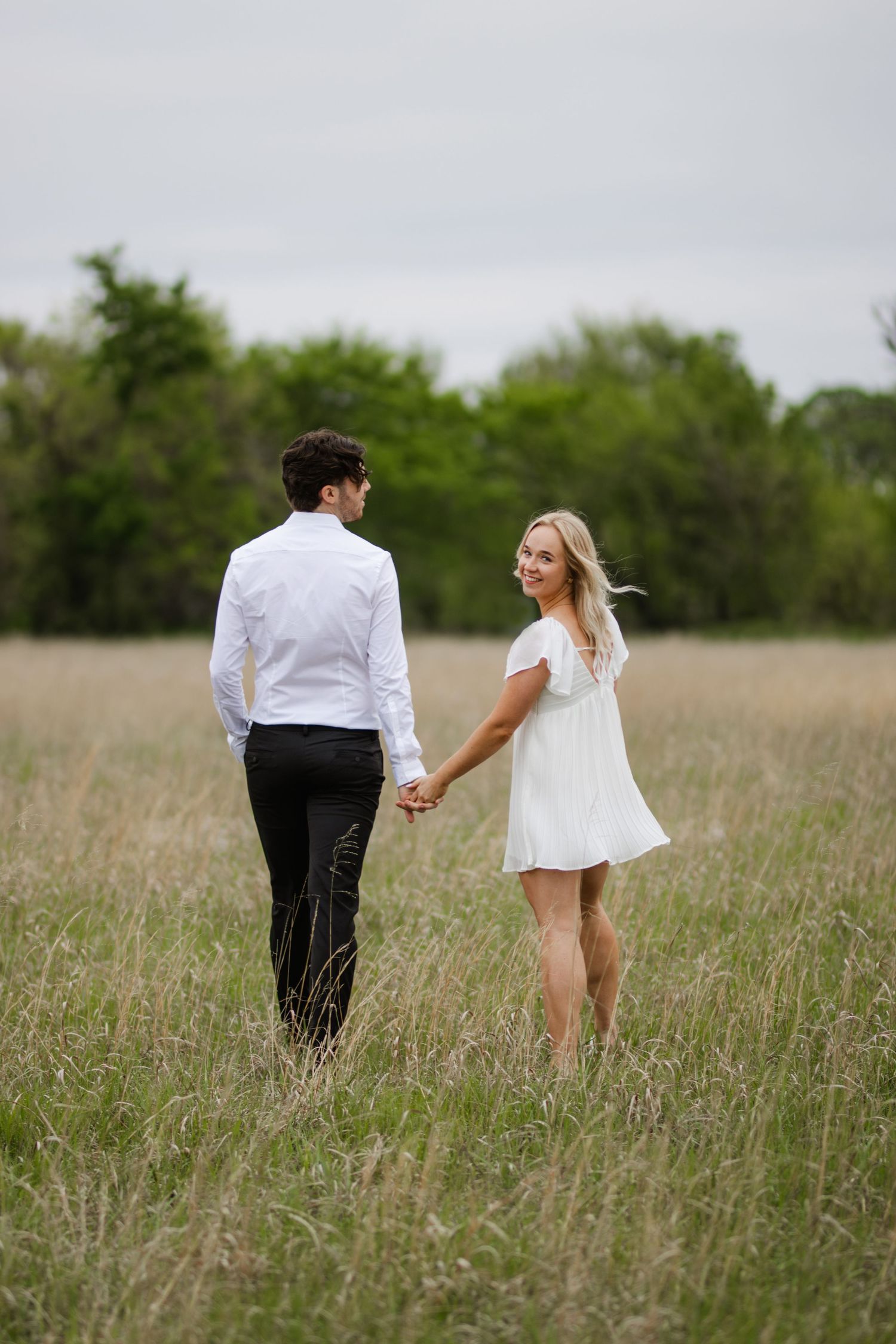 Couple holds hands while standing in a tall grass field on an overcast day, wearing white and black formal attire.