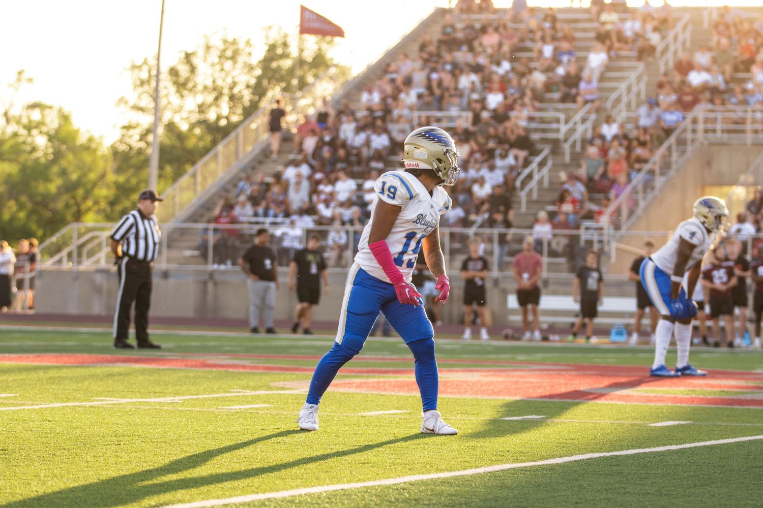 Football player in blue and white uniform stands ready on field during sunset game.