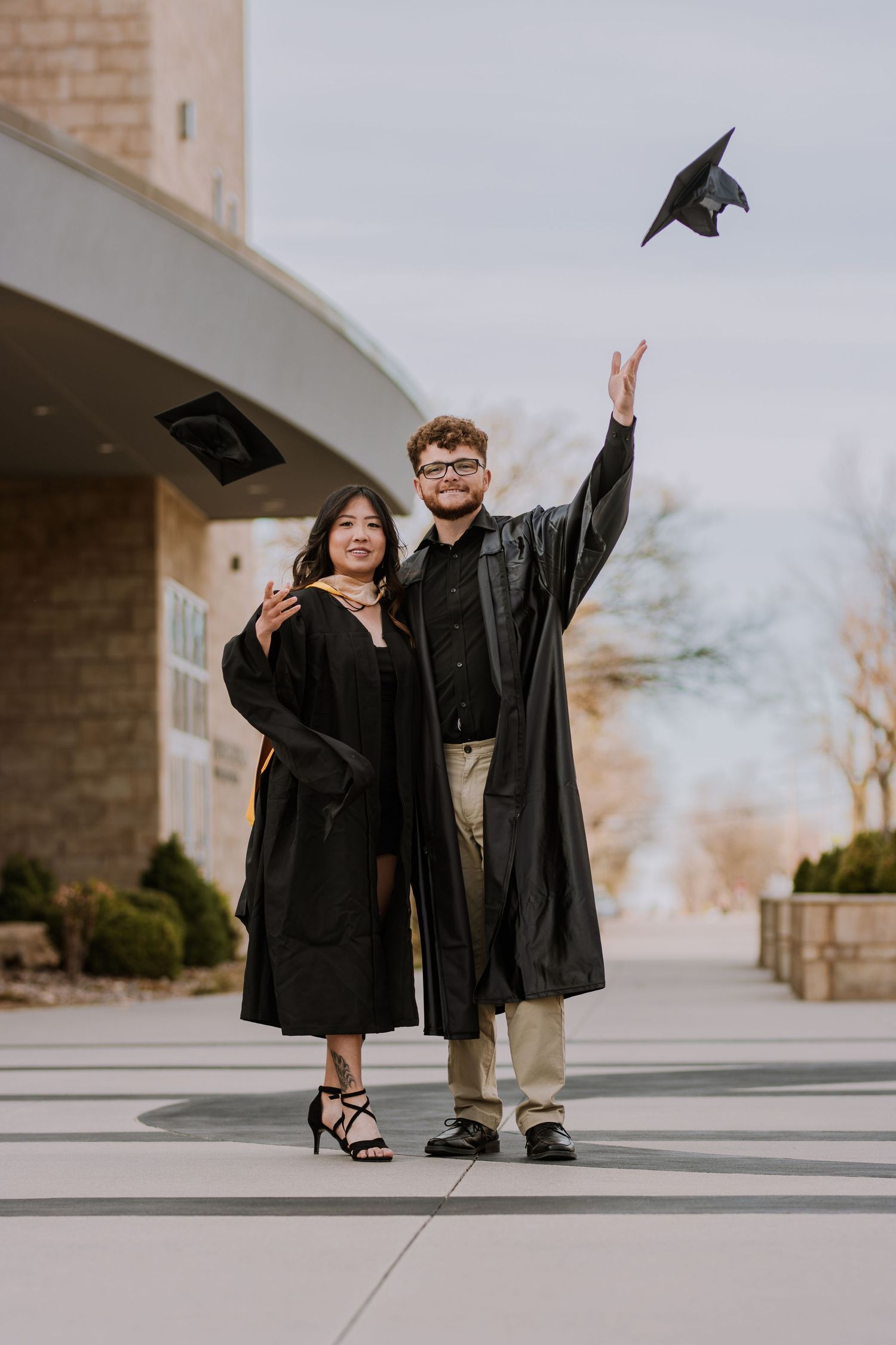 Graduation photo of two people in black caps and gowns throwing mortarboard in the air.