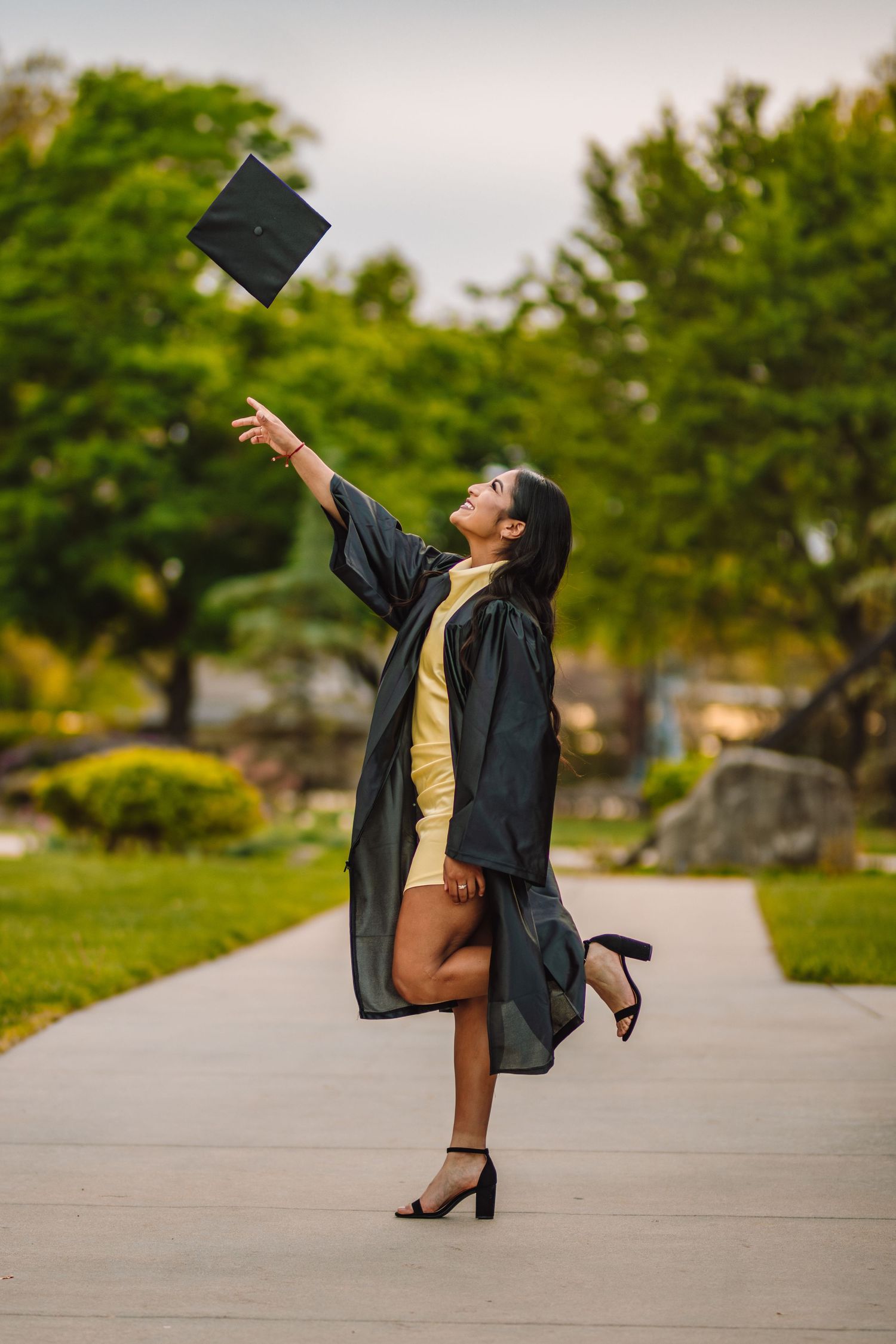 Joyful graduate in green cap and gown throws their graduation cap in the air while standing on a campus pathway.