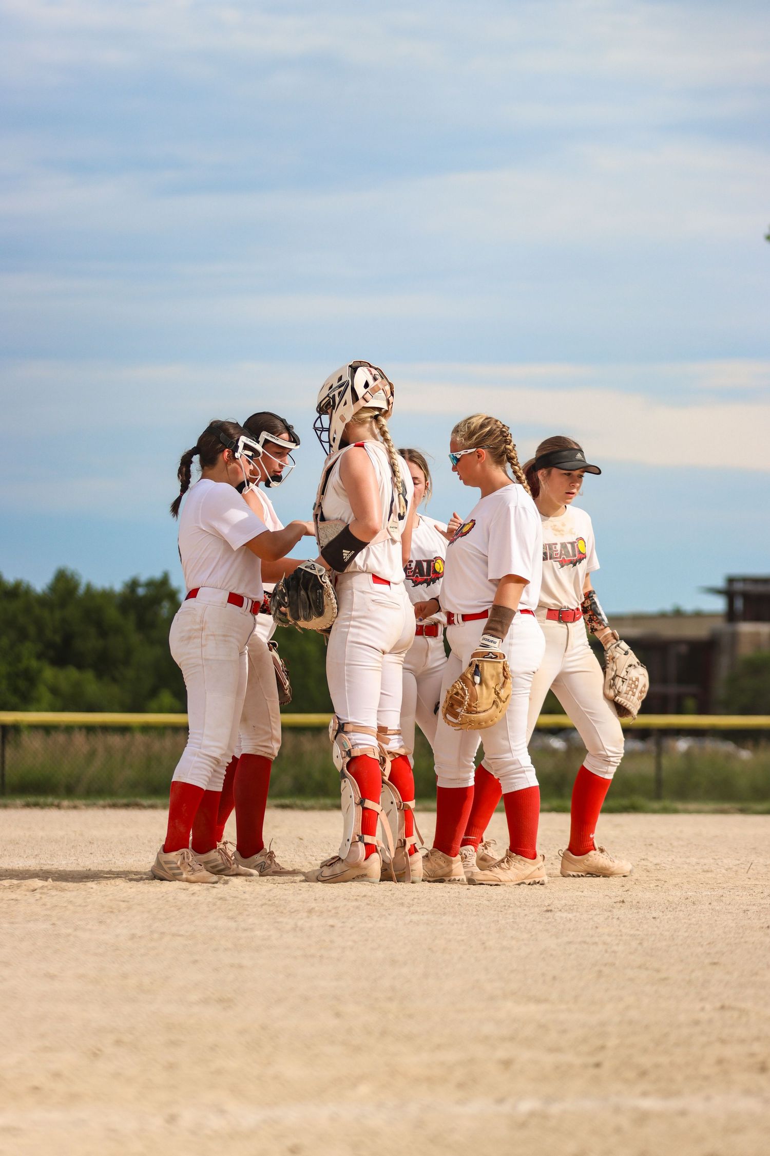 Team of softball players in white uniforms huddle together on dirt field during game.