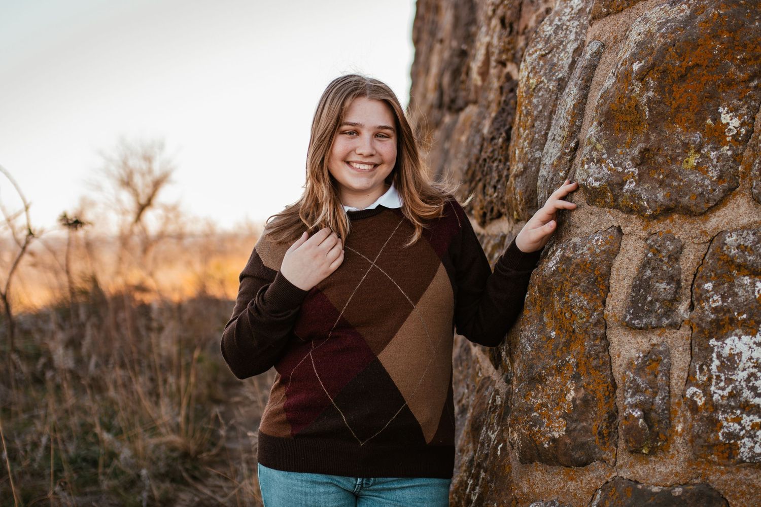 Person wearing brown sweater poses next to stone wall in autumn landscape.