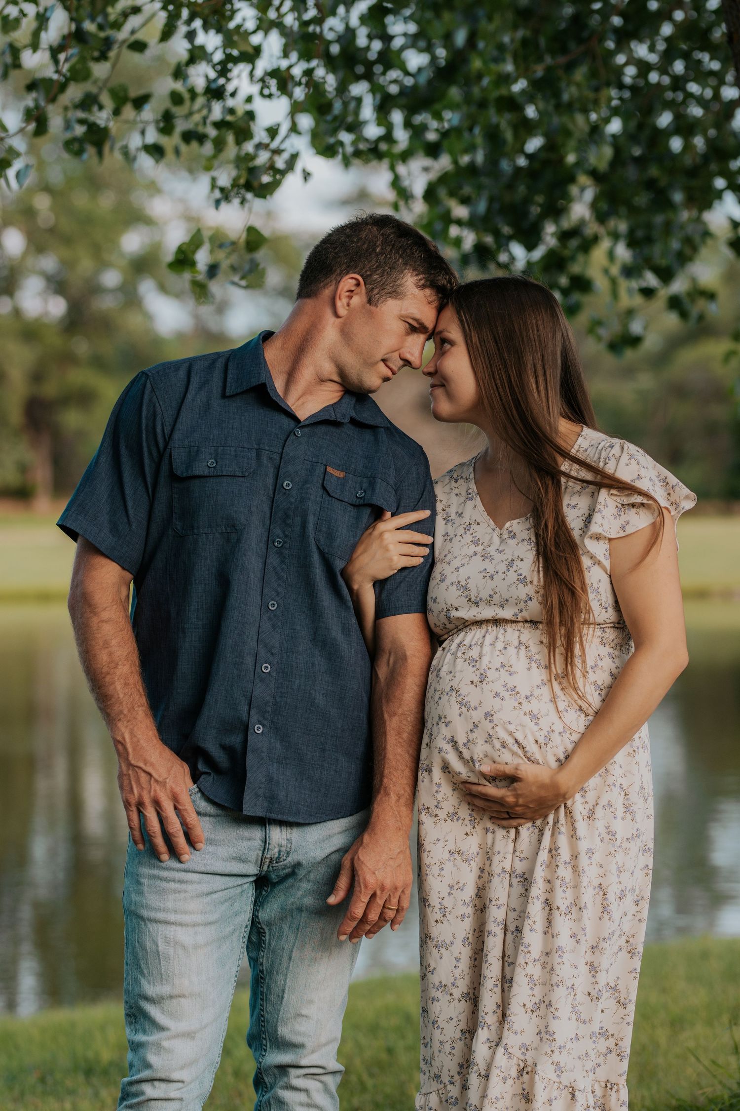 A romantic couple poses together outdoors by a lake, with her wearing a lace dress and him in a navy shirt and jeans.