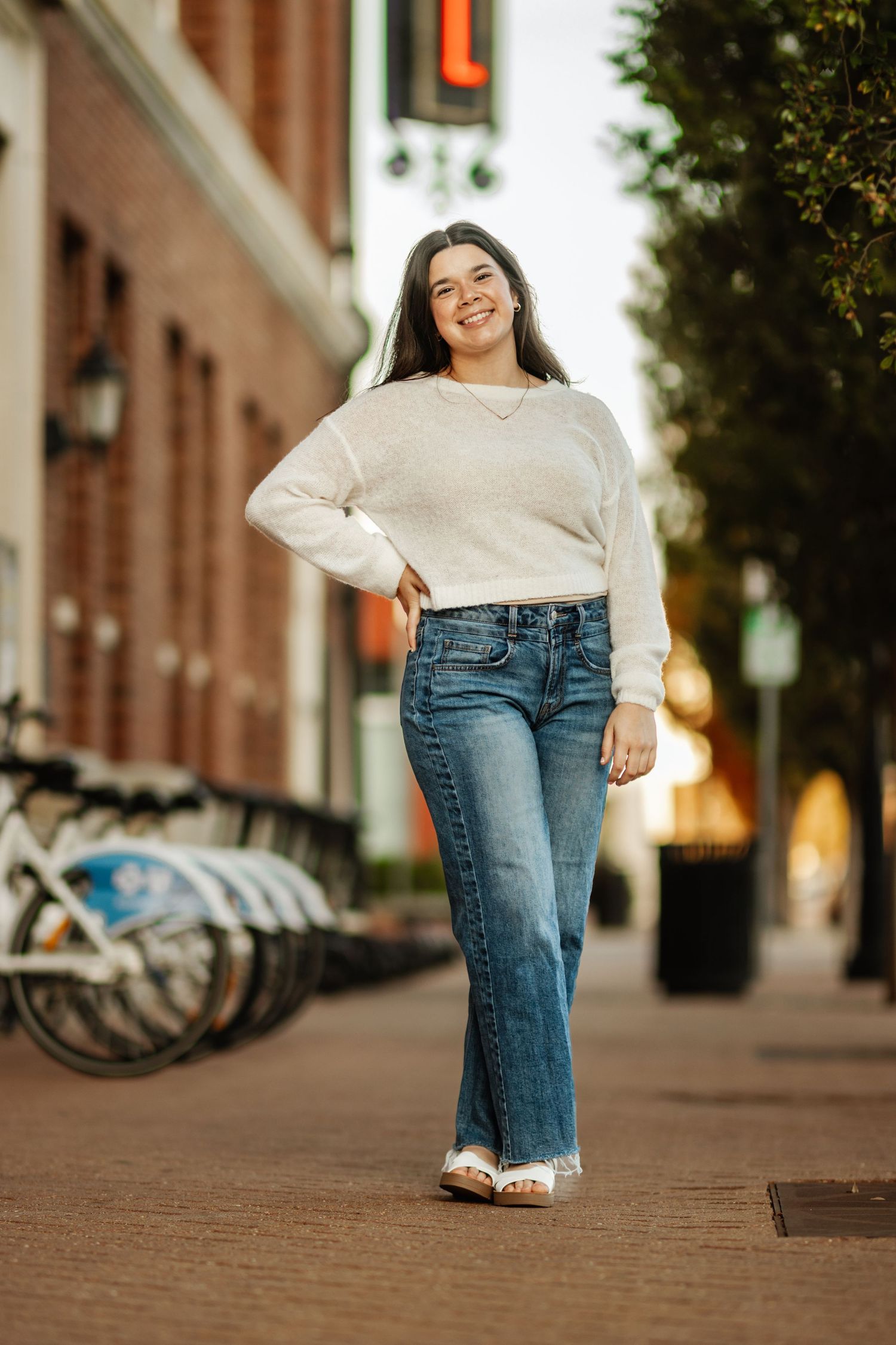 Casual street style outfit with cream sweater and flared jeans on urban sidewalk with bikes in background.