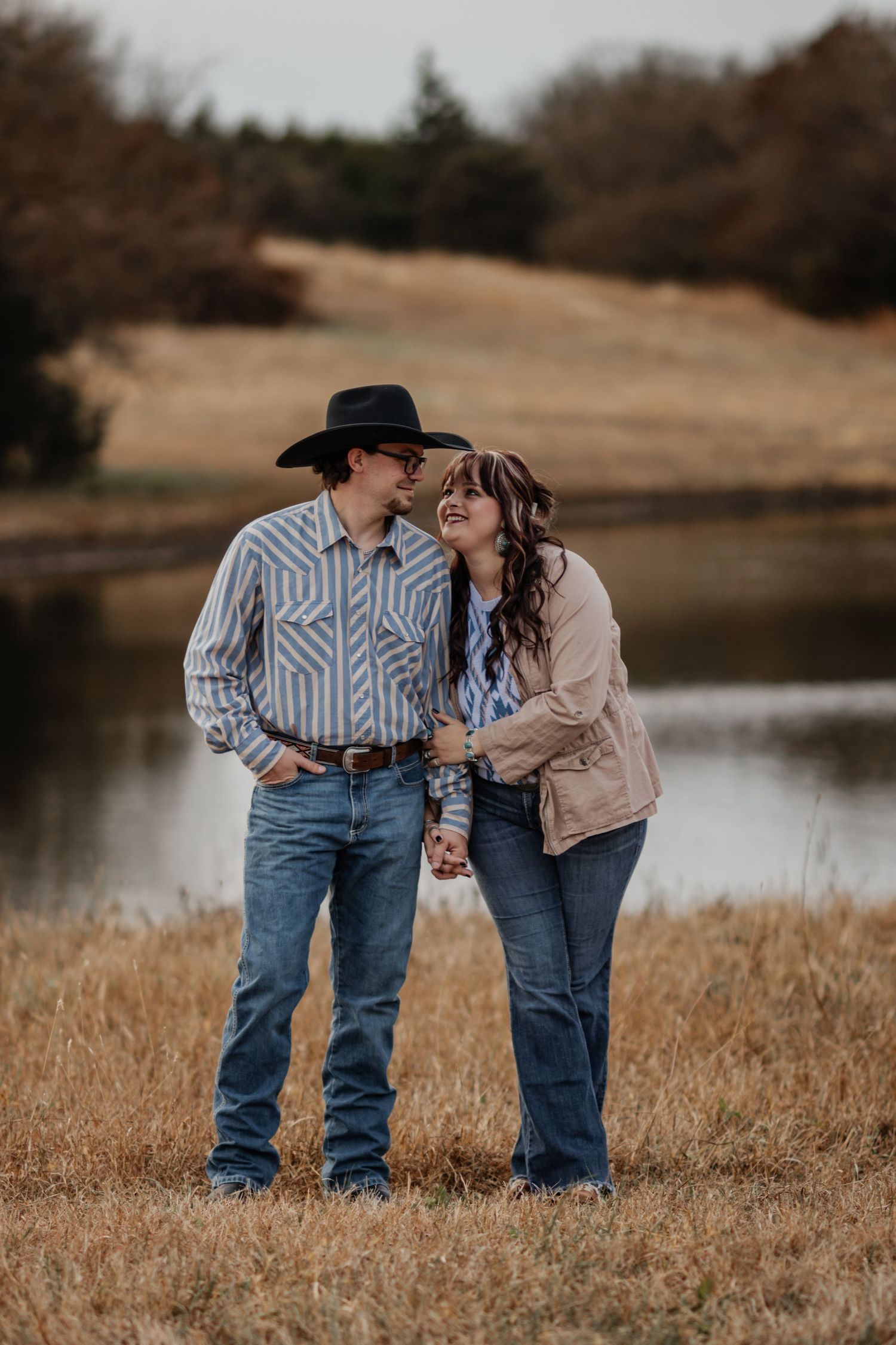 A couple in western attire shares an intimate moment by a serene lake surrounded by golden grass at sunset.