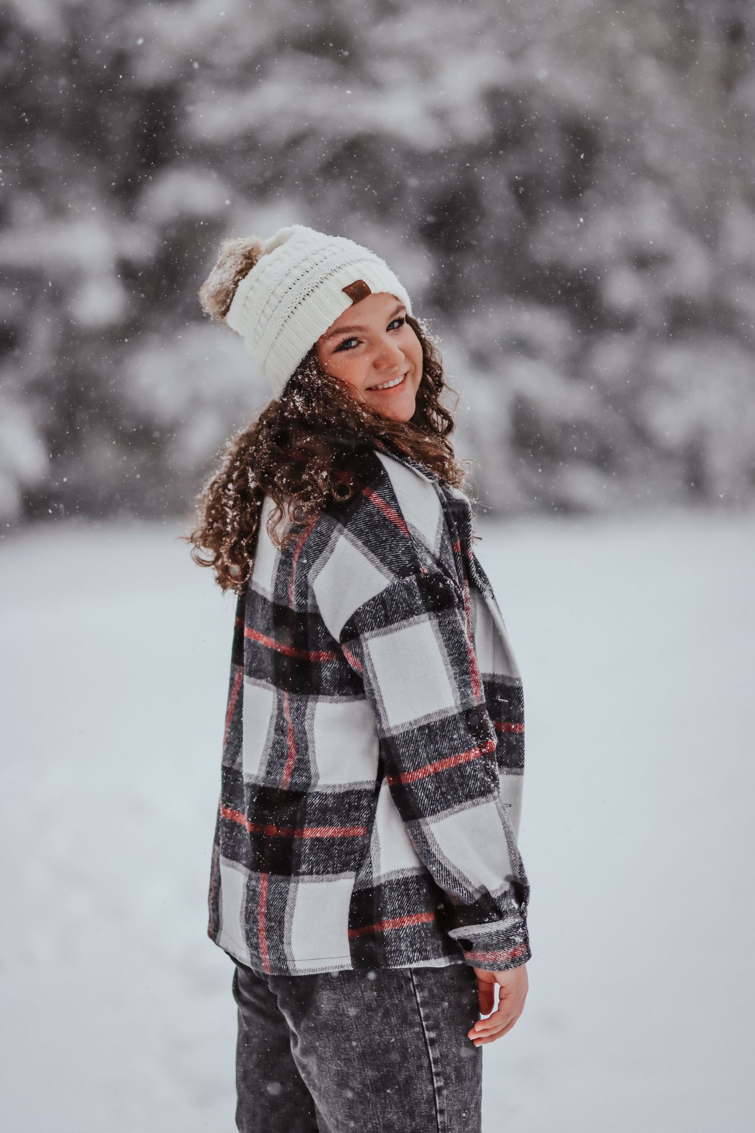 Person wearing plaid coat and beanie smiling during snowy winter photoshoot.
