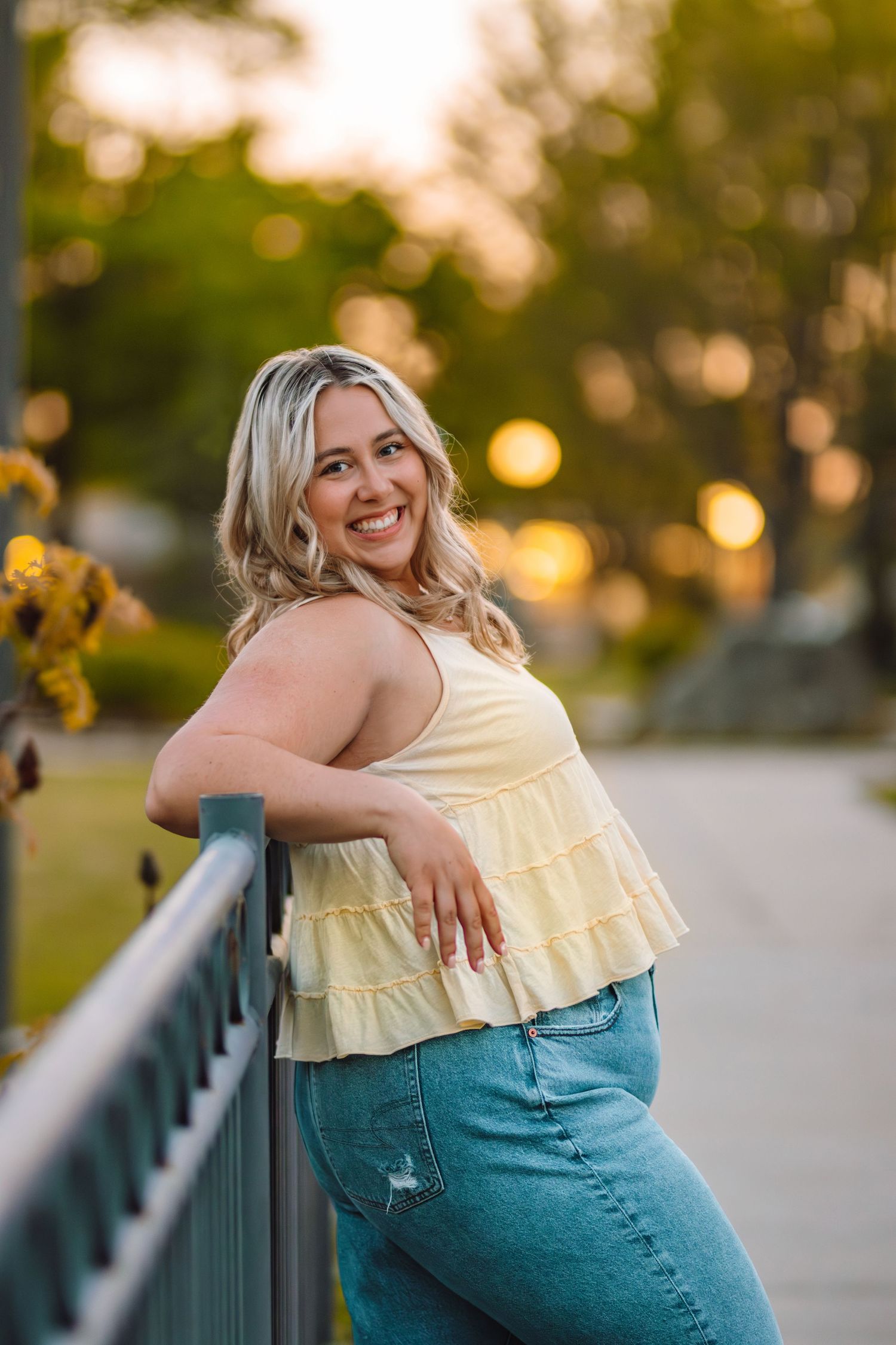Woman in yellow ruffled top leans against railing during golden hour outdoor portrait session.