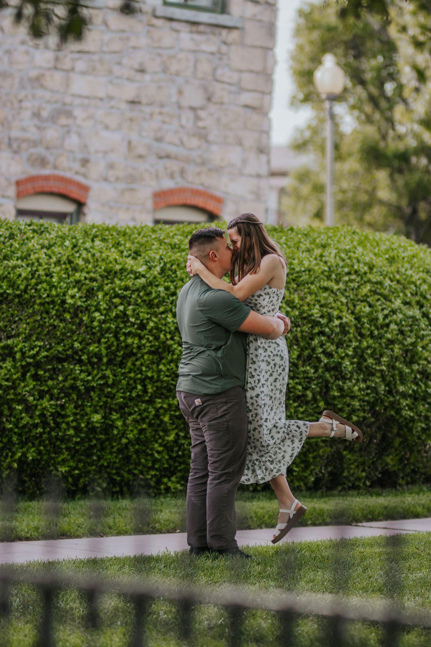 A couple shares a romantic embrace in front of stone wall and green hedges at golden hour.