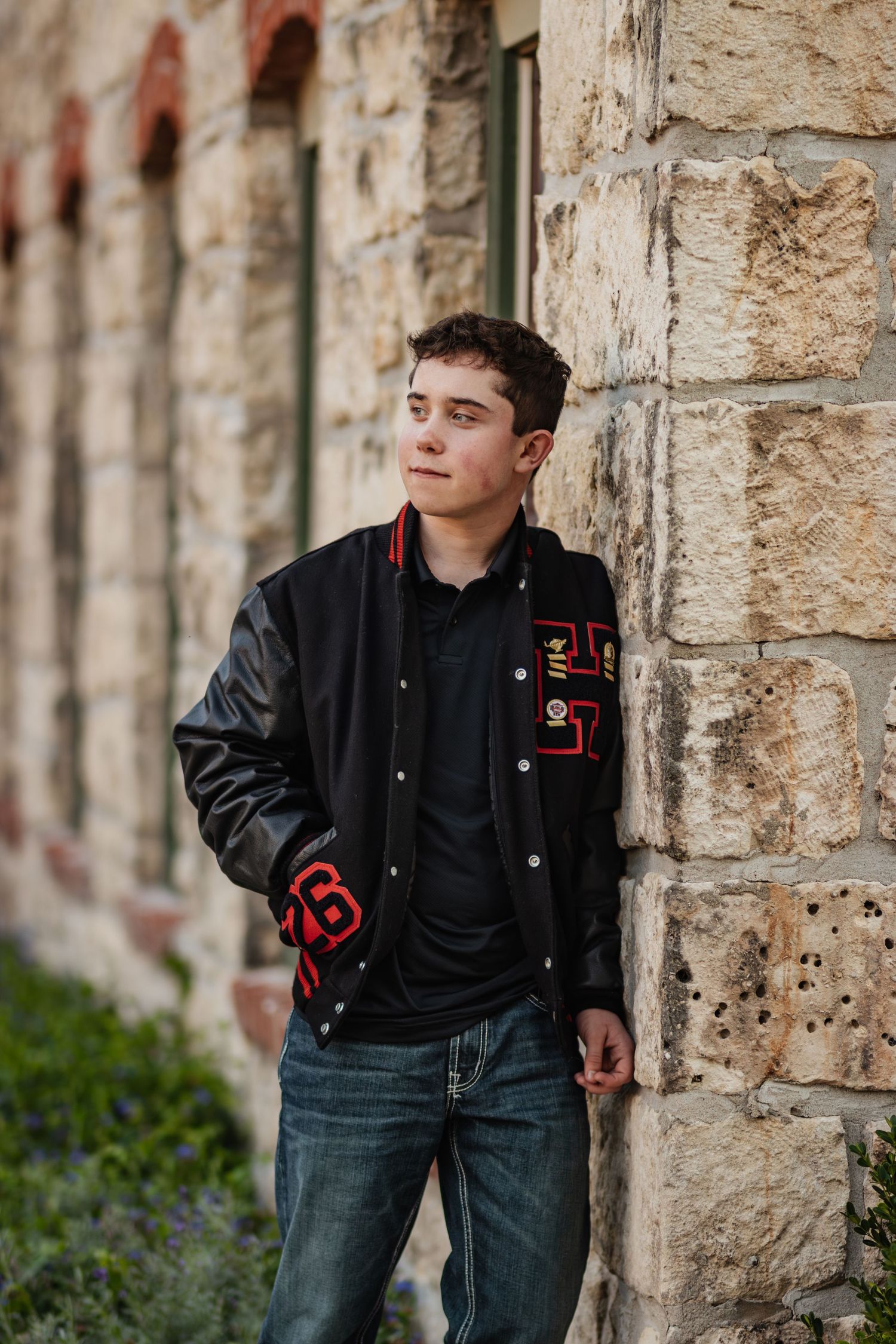 A young person in a black varsity jacket leans against a stone wall while looking away from the camera.