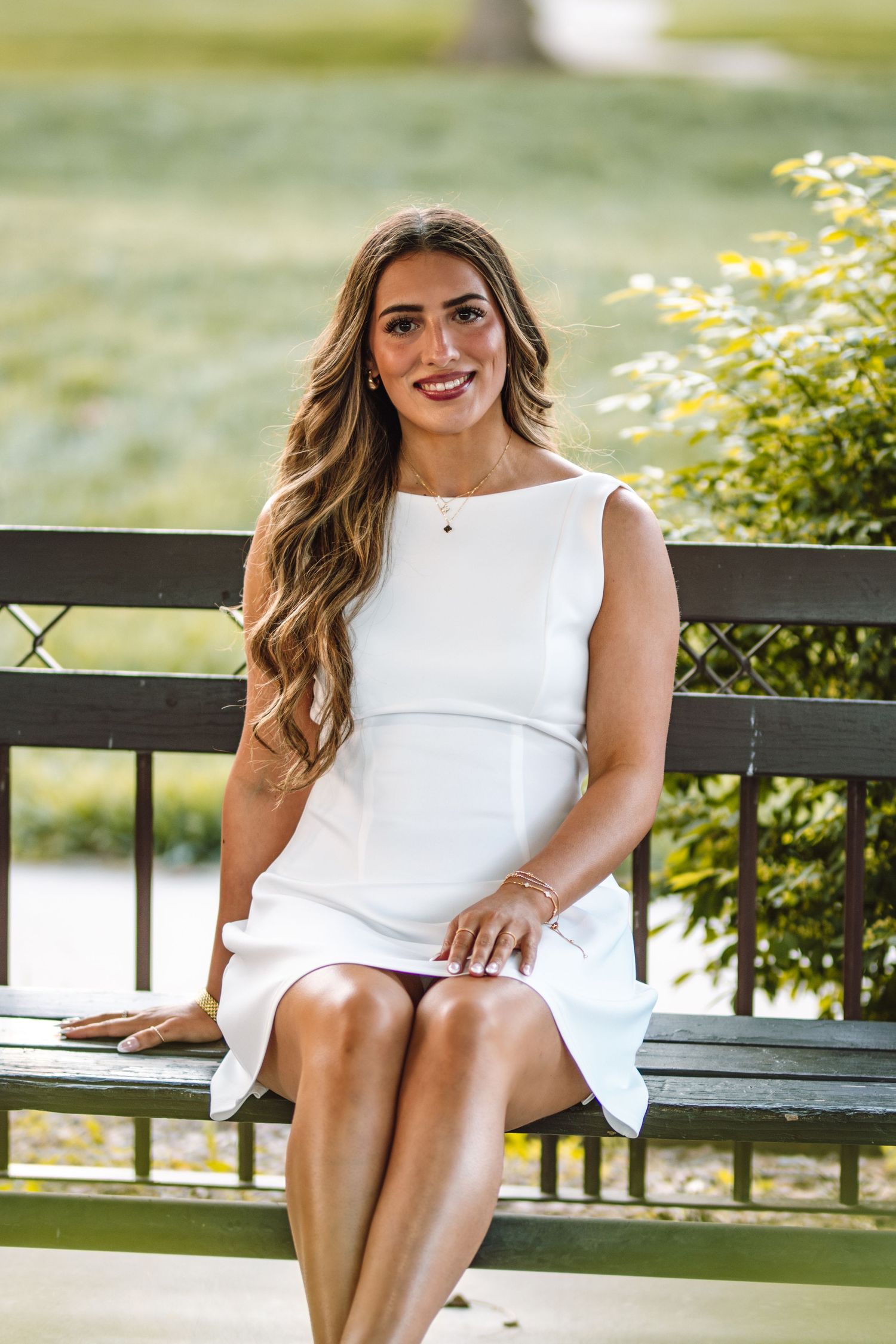 A woman in an elegant sleeveless white dress sits on a park bench on a bright summer day.