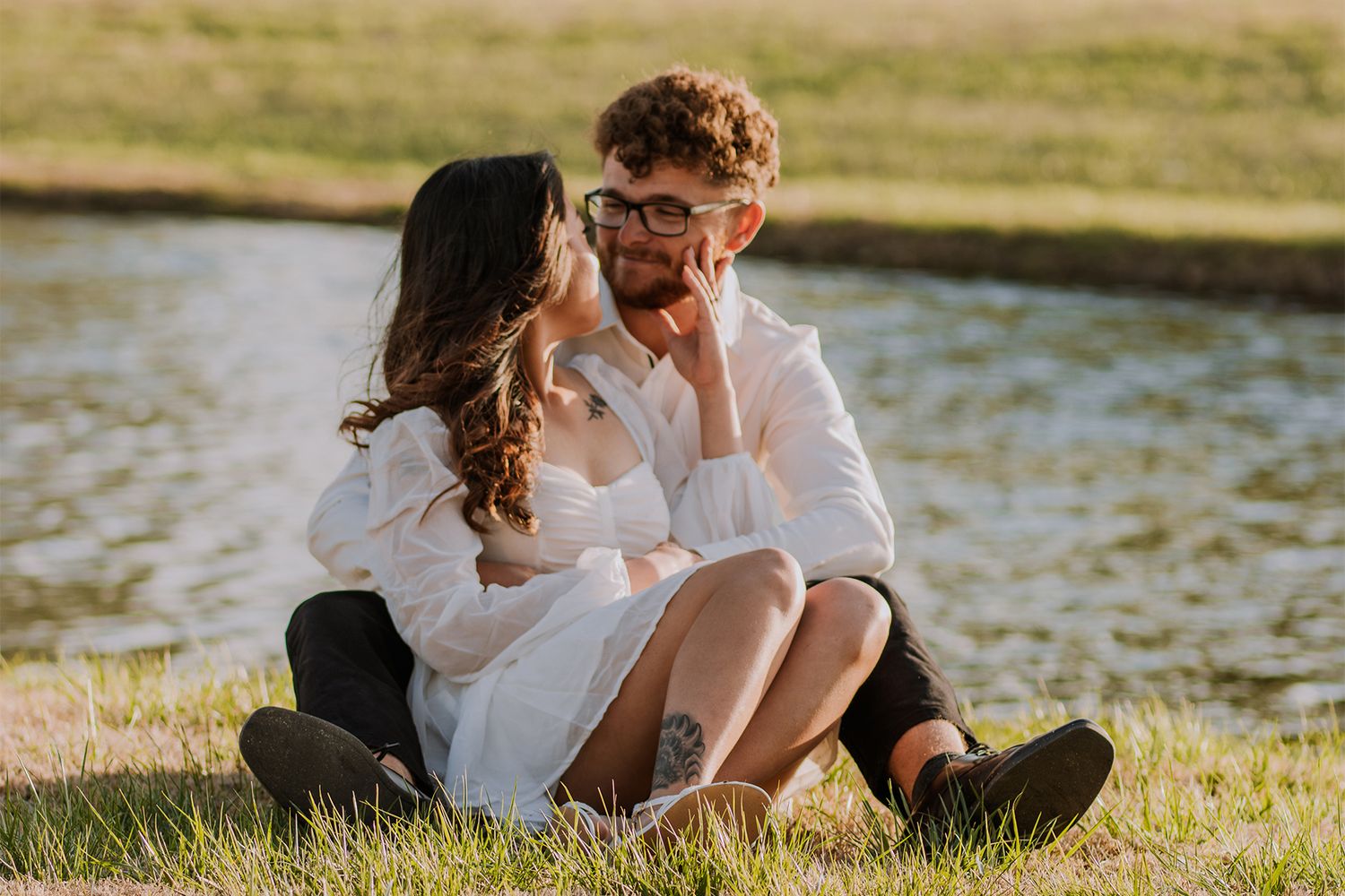 Couple in white clothes sitting together on grass by water during sunset.