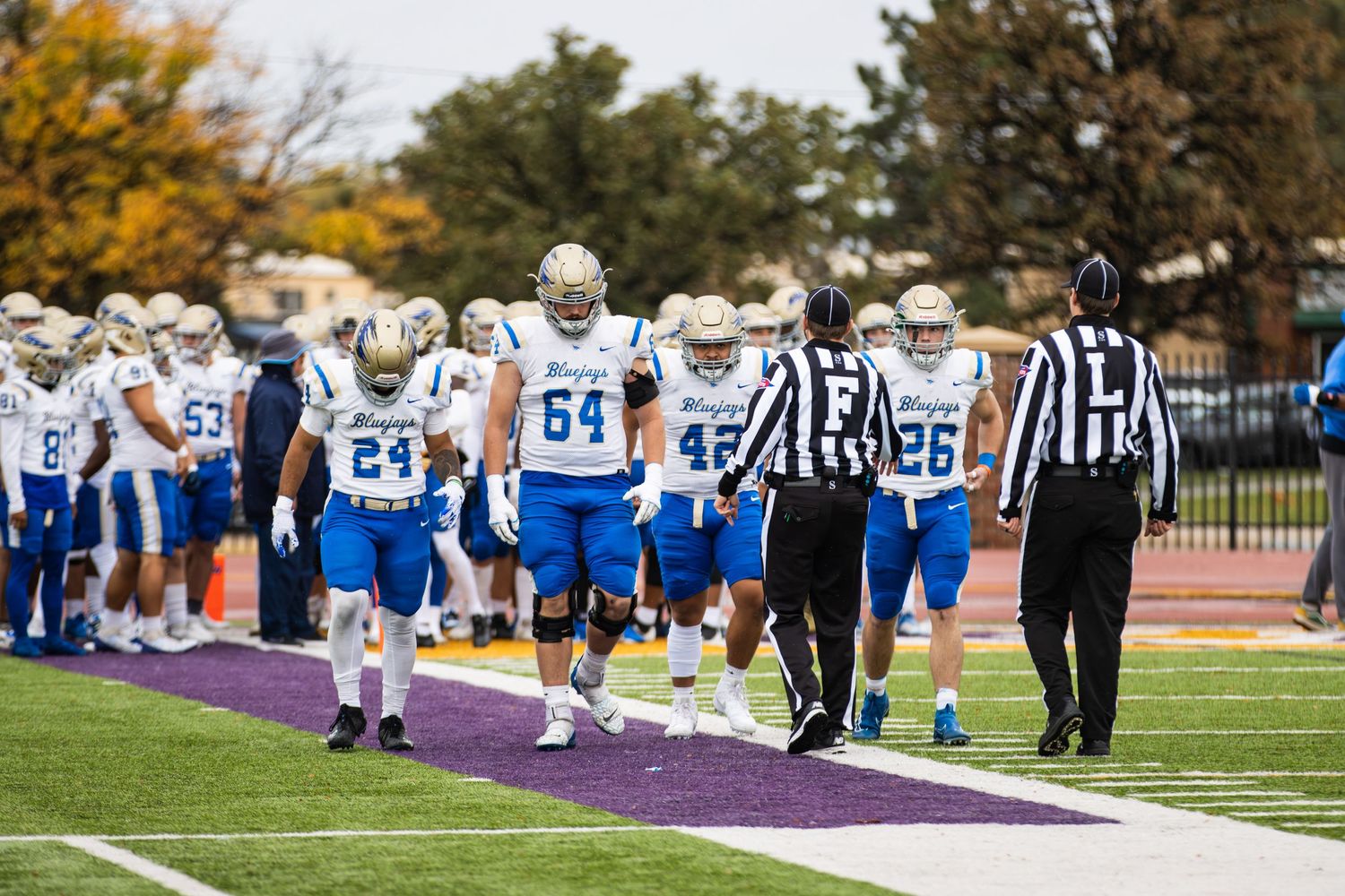 Football referees and players in blue uniforms walking along sideline during game.