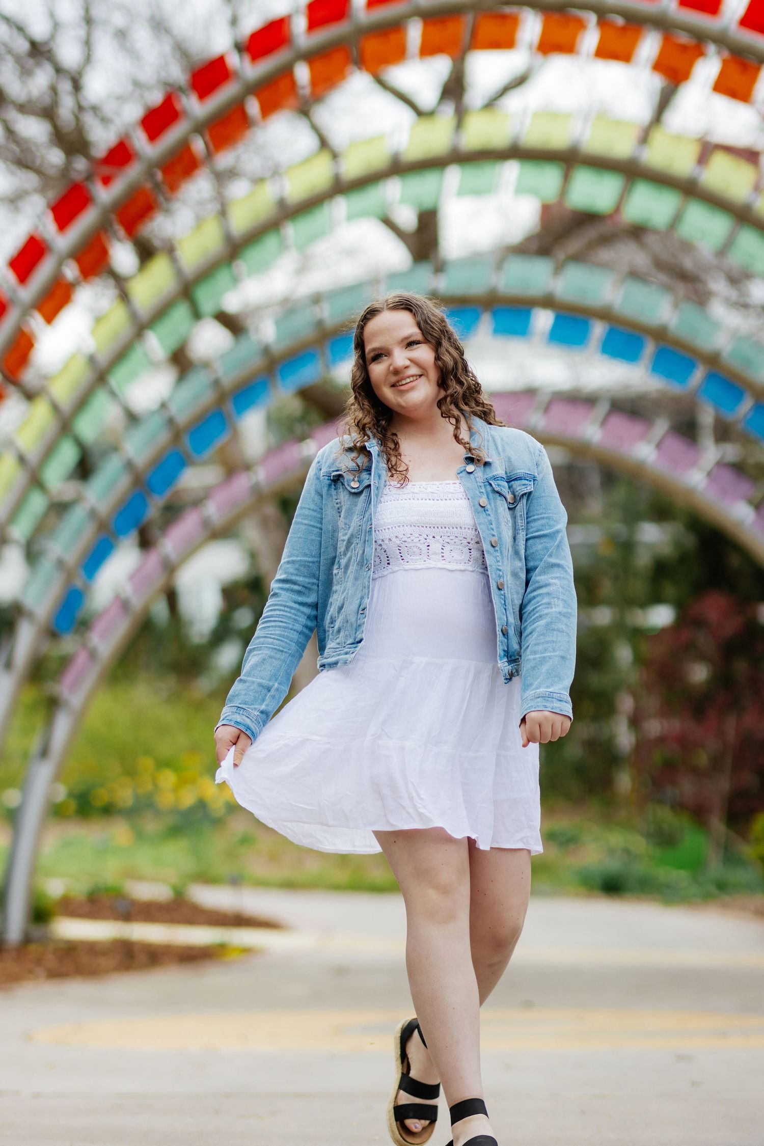 A woman in white dress and denim jacket poses under a colorful rainbow arch outdoors.