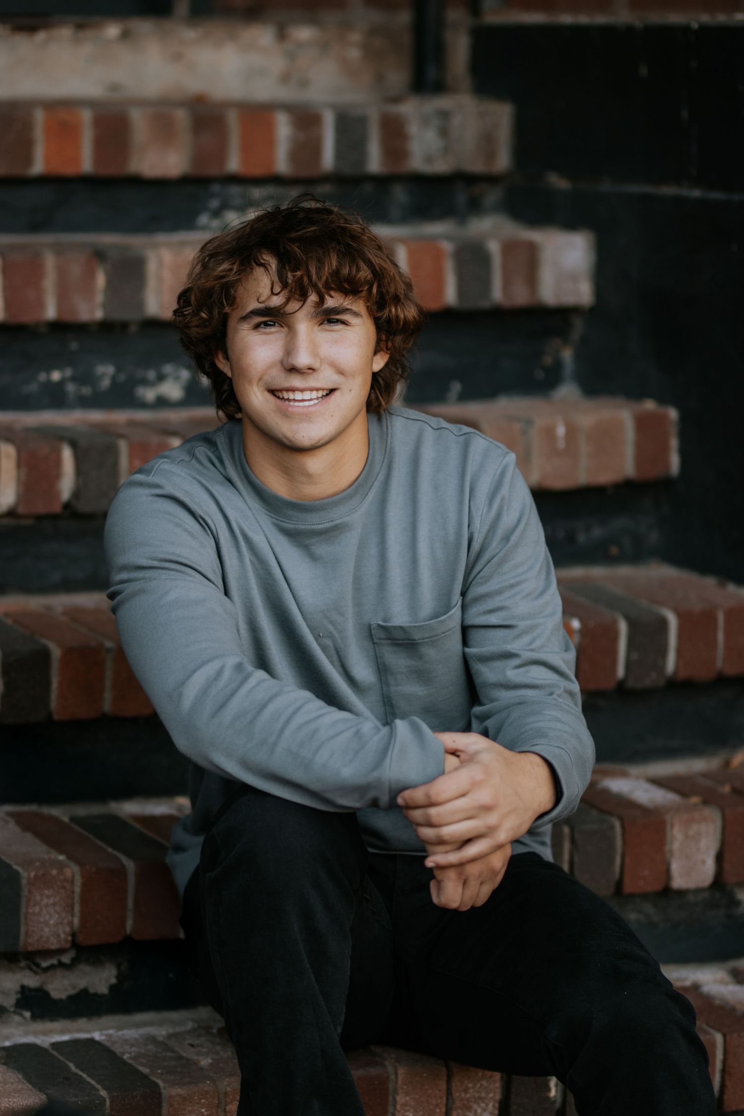 Young person in gray sweatshirt sitting on brick steps smiling at camera during outdoor portrait session.