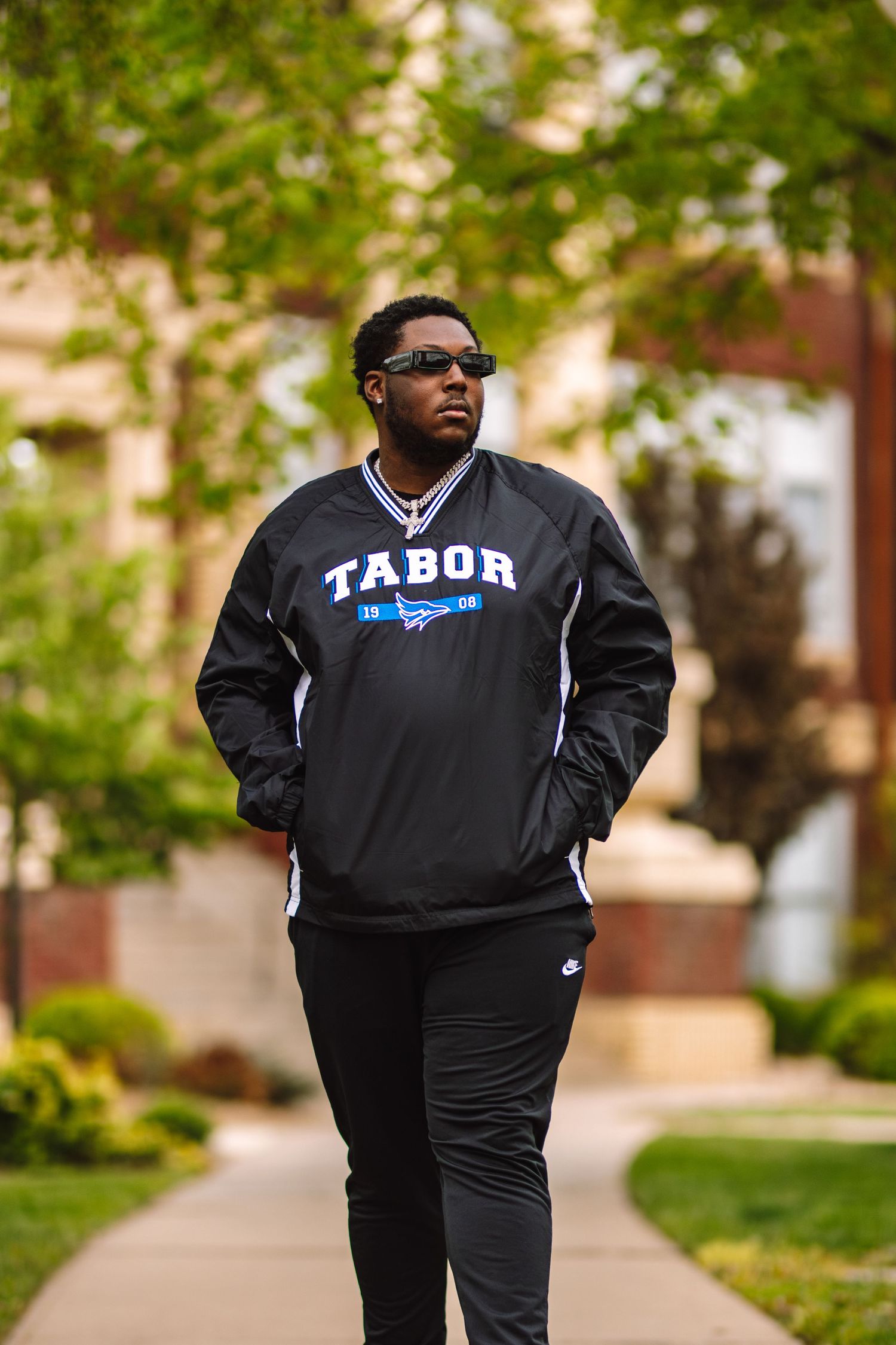 Three photos of someone wearing a black tracksuit and sunglasses walking on a paved path with trees and buildings in background.