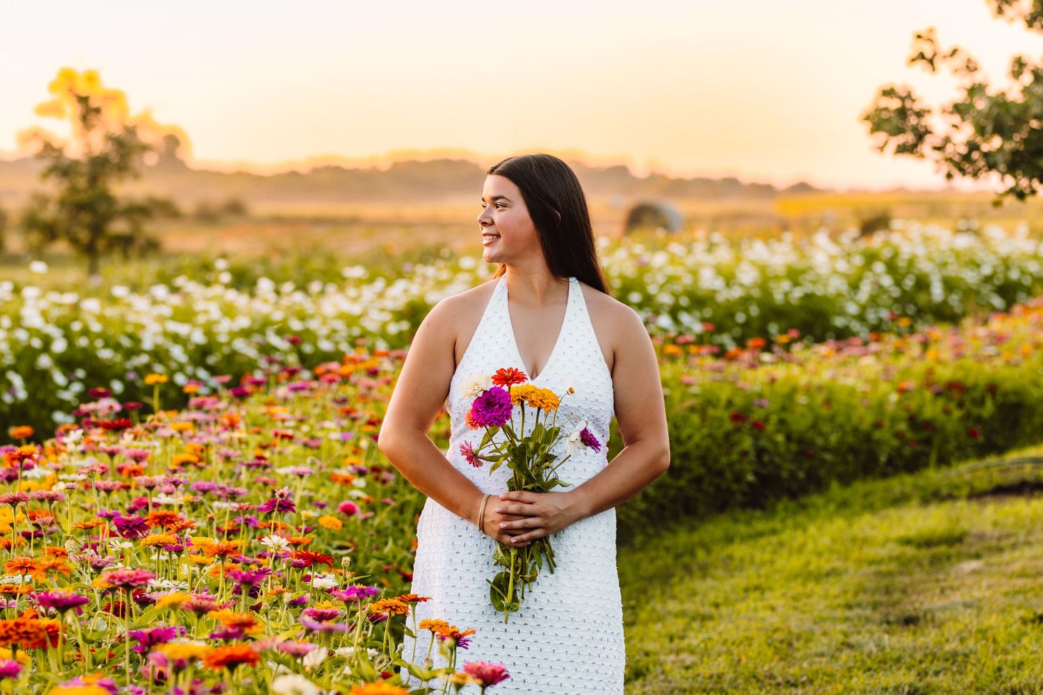 A woman in a white dress sits joyfully in a vibrant flower field during golden hour sunset.