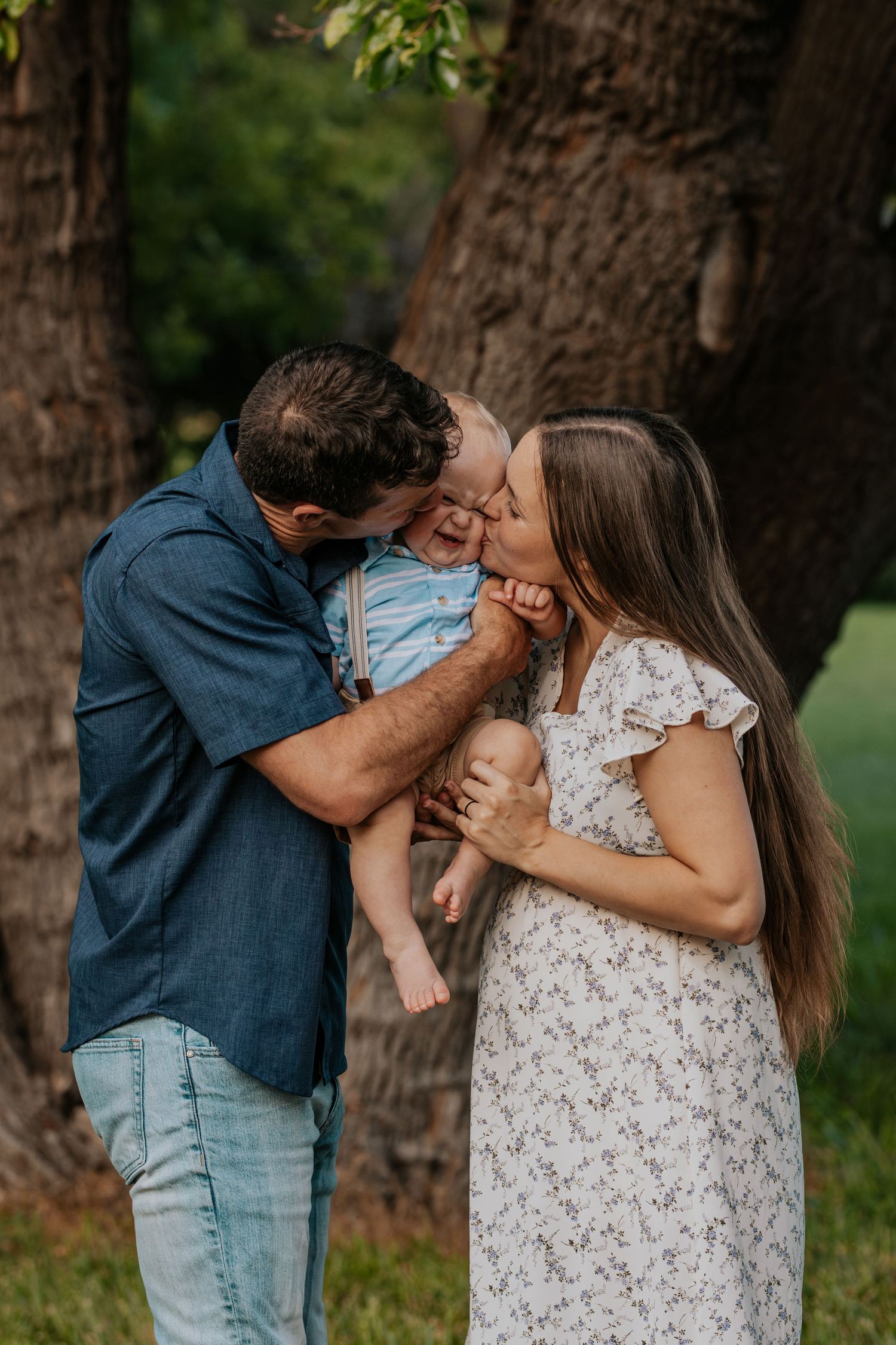 An affectionate couple shares tender kisses against a tree trunk background in a natural outdoor setting.