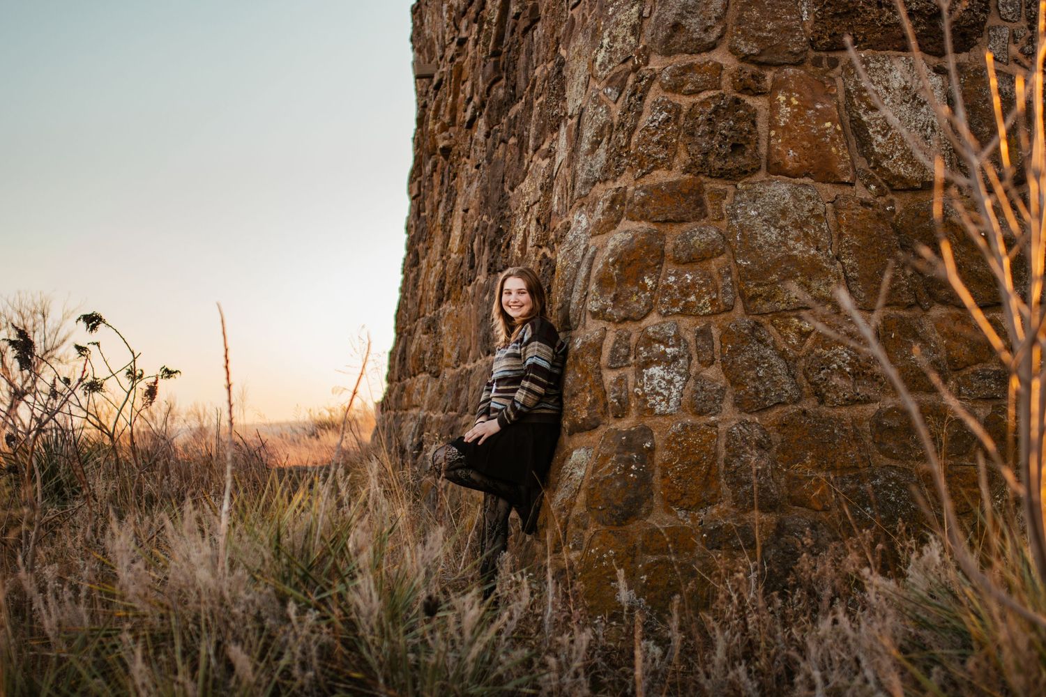 Person in dark coat leaning against stone wall during golden hour.
