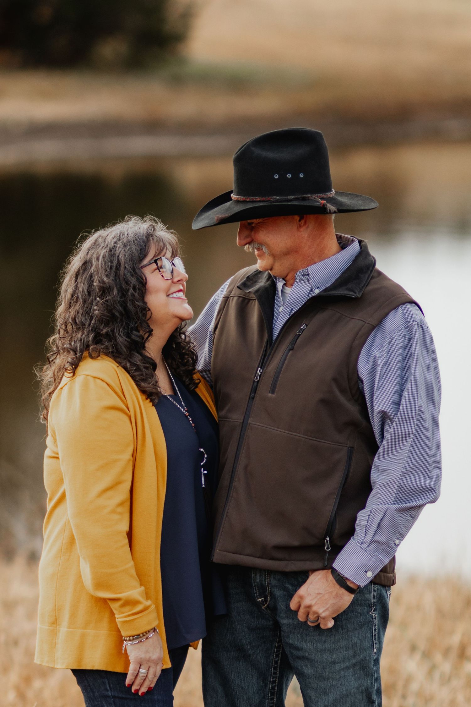 A couple in western attire shares a tender moment by a lake with the woman wearing yellow and man in cowboy hat.