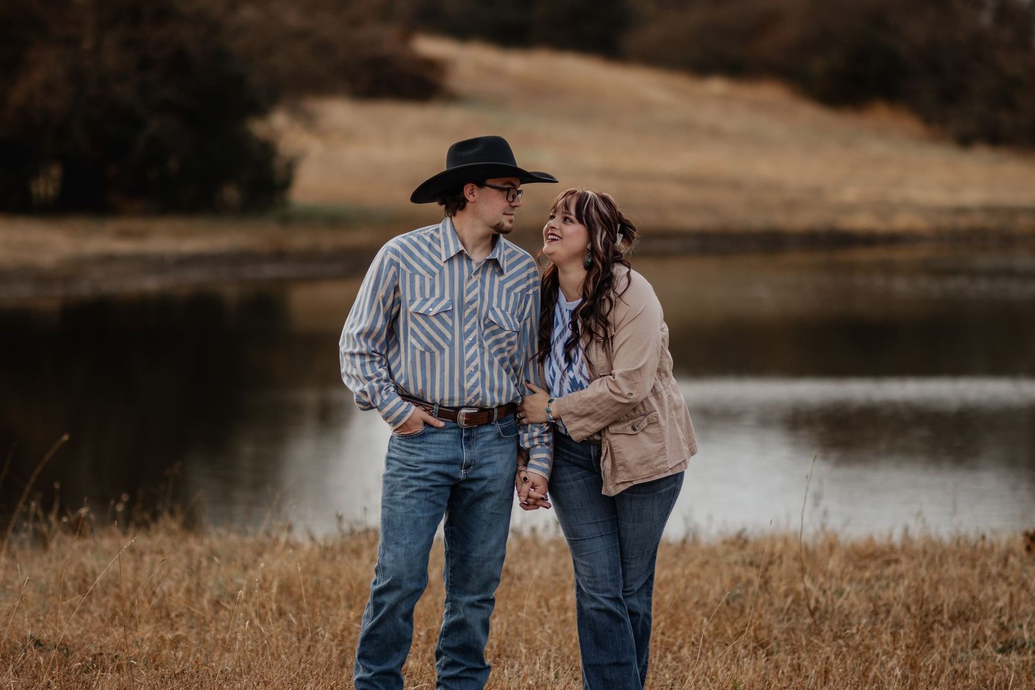 A couple in western attire shares an intimate moment by a serene lake during golden hour.