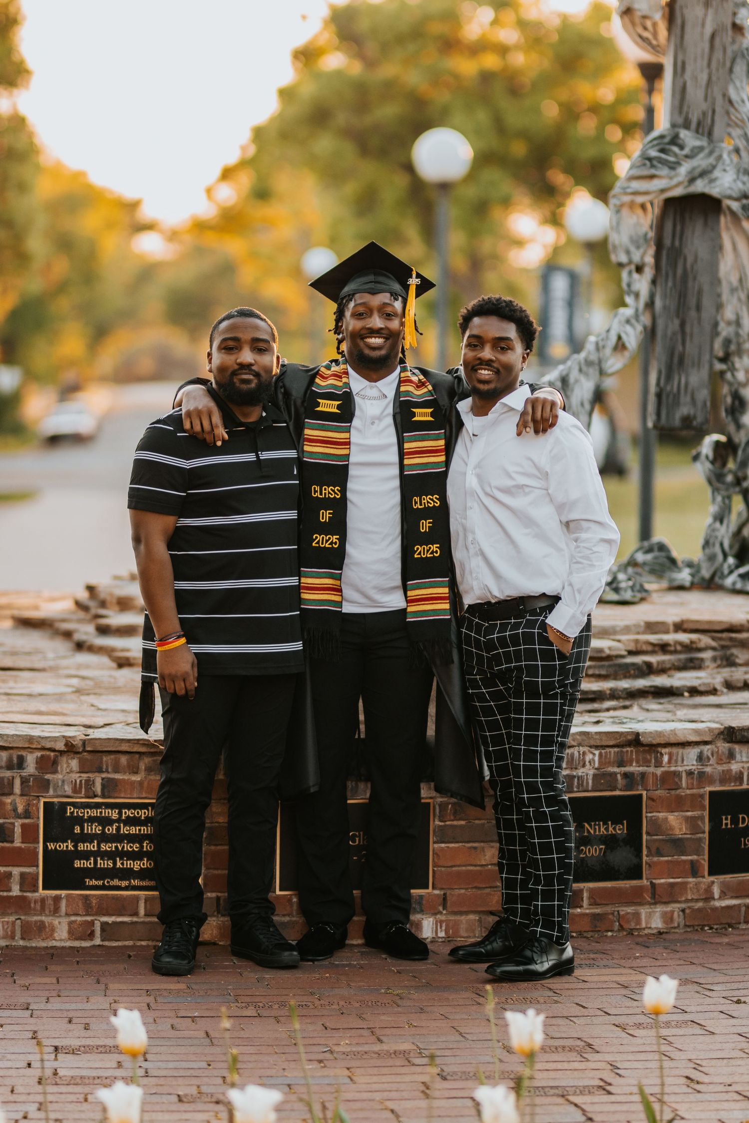 A graduation celebration on a brick sidewalk during autumn with the graduate wearing ceremonial stoles and cap.