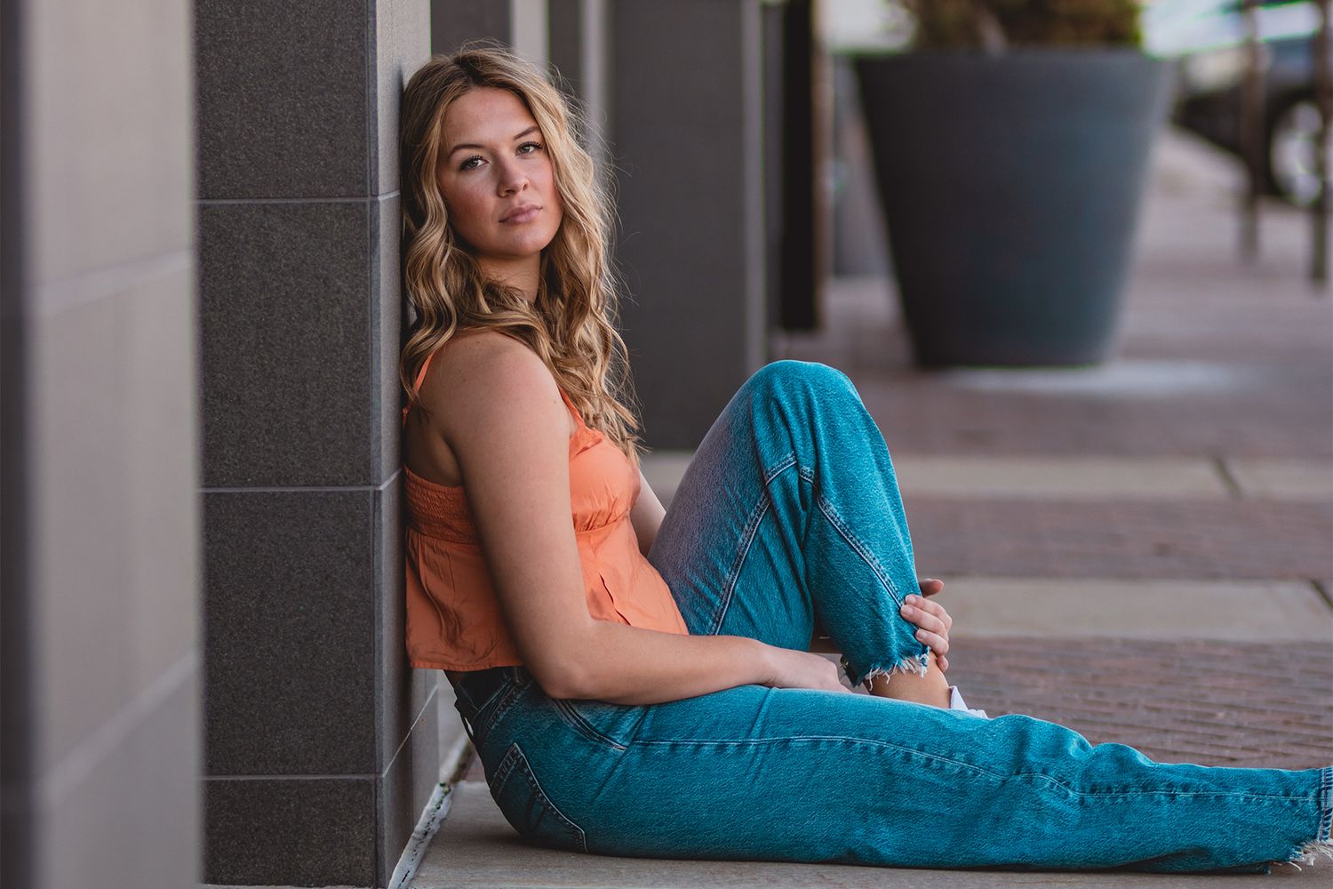 Casual portrait of someone in orange top and blue jeans sitting against a gray wall.