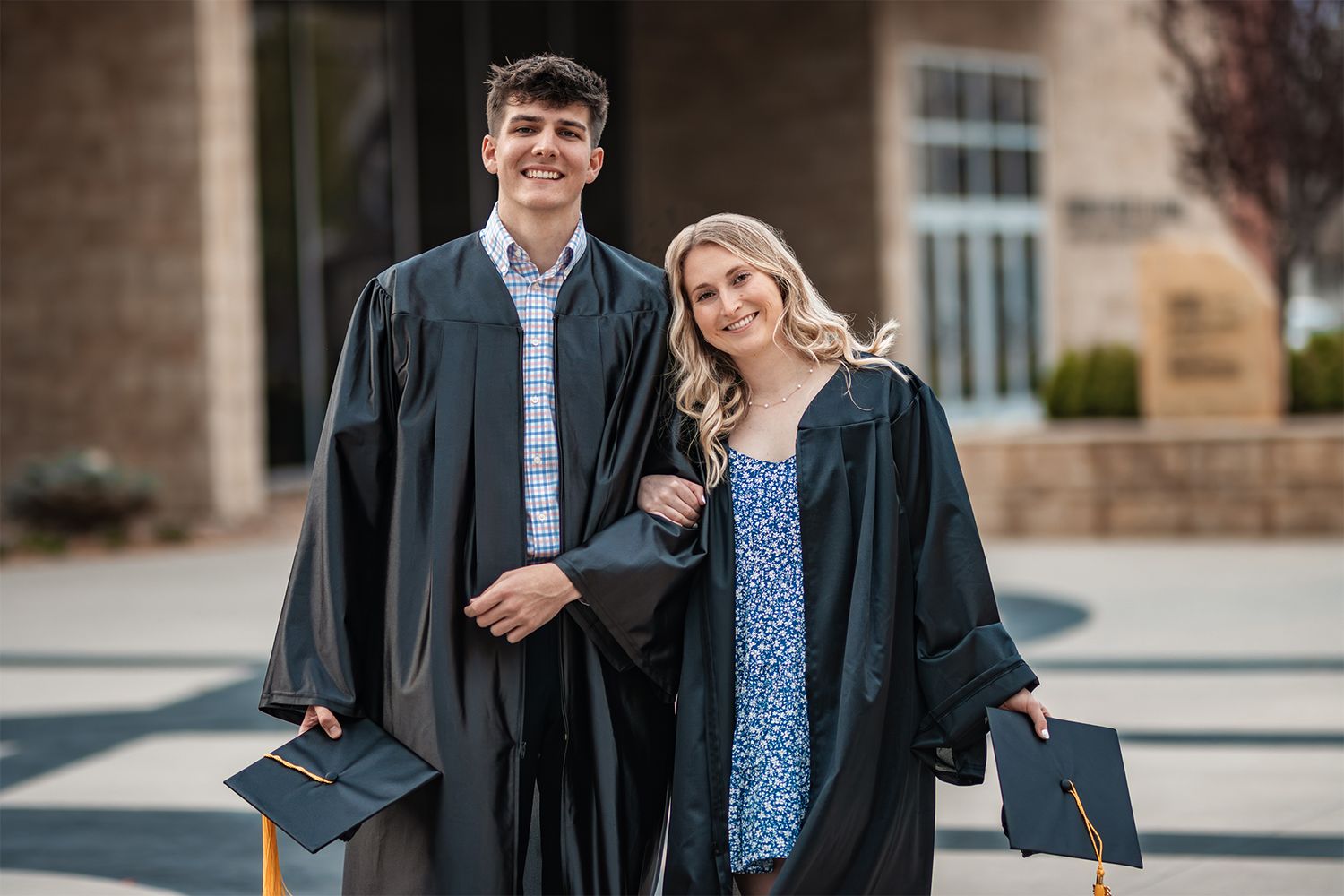 Graduates wearing black academic regalia smile together while holding their diplomas outdoors.