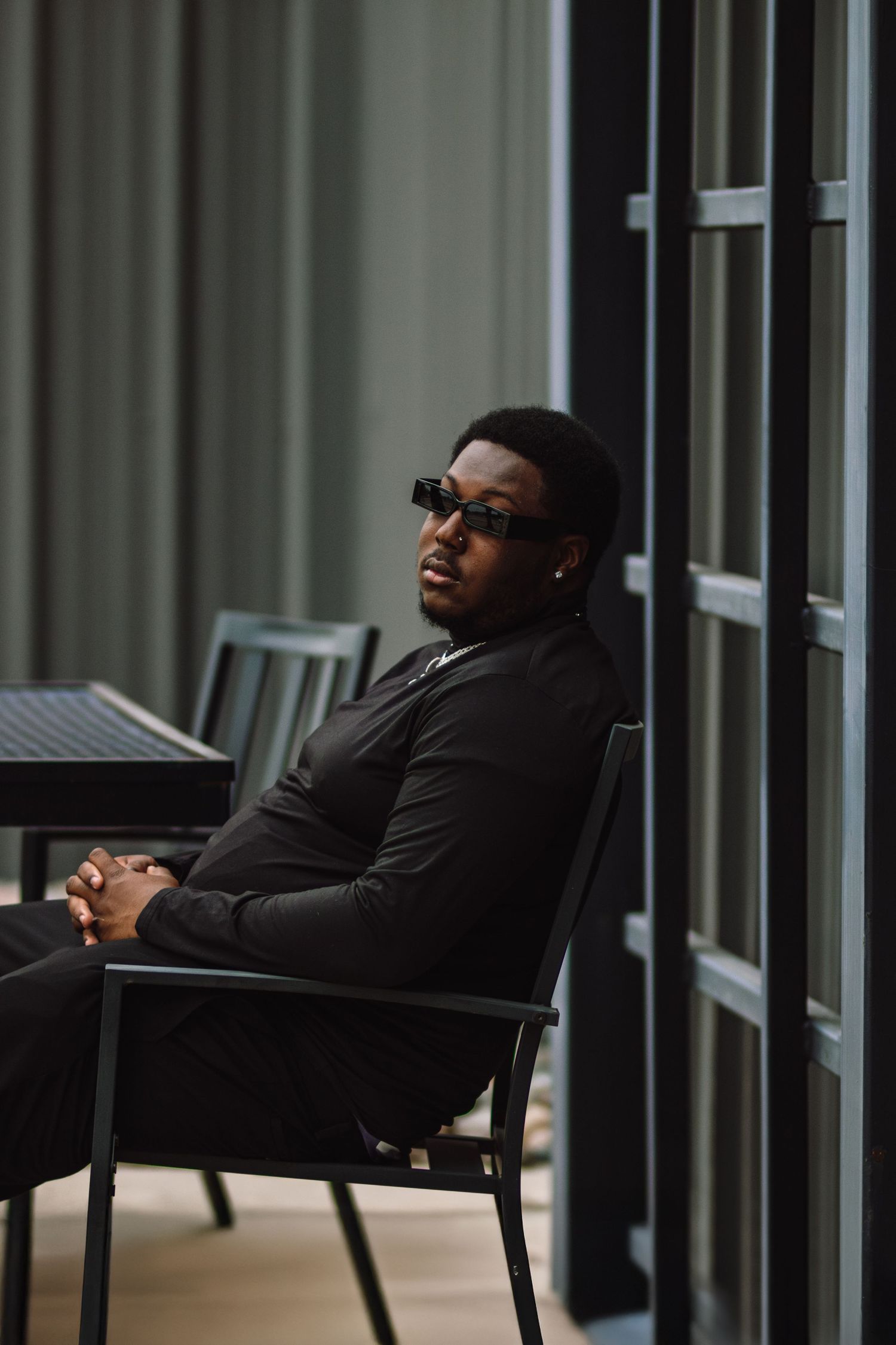 A person in glasses reclines in a black chair against a backdrop of vertical lines and windows.