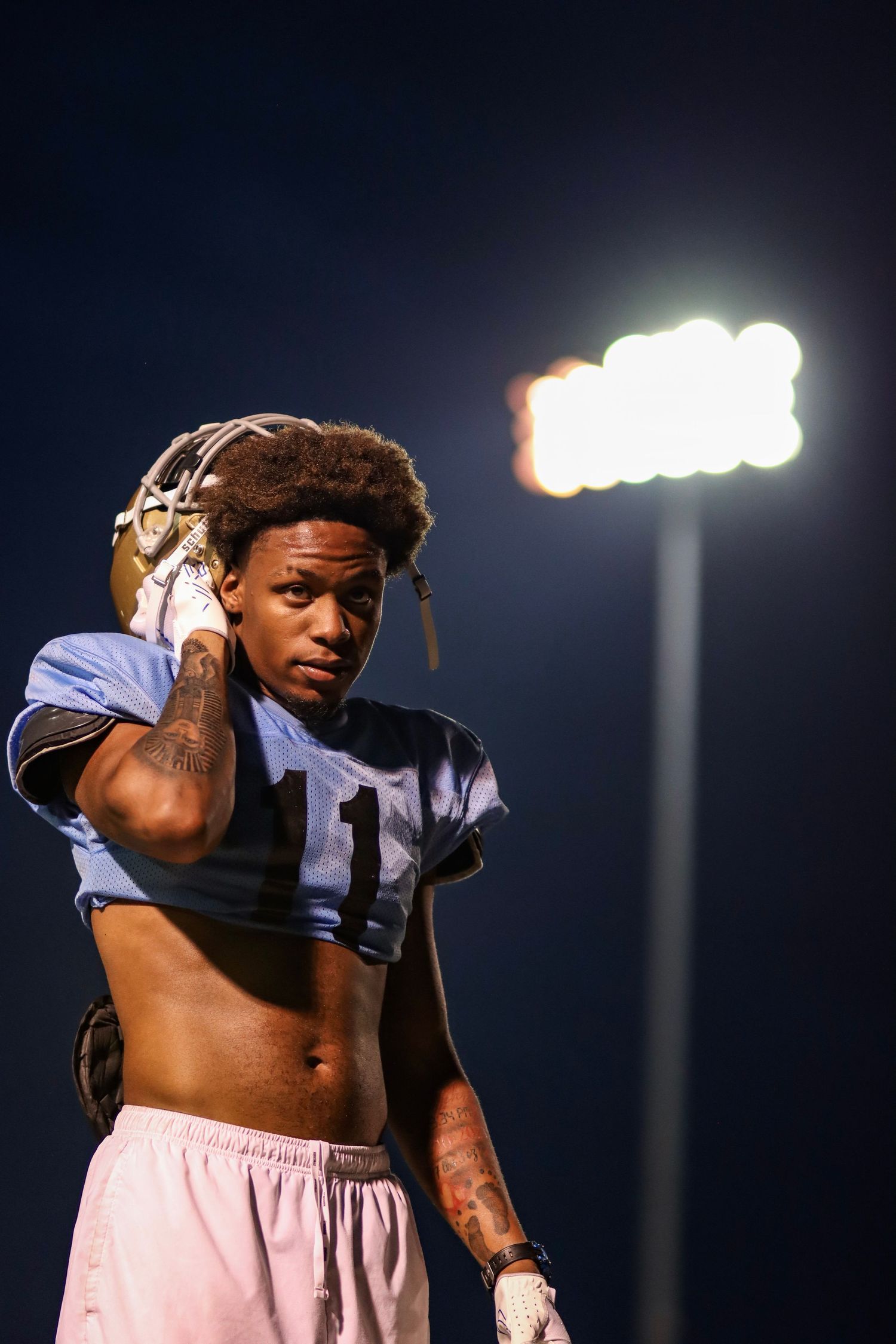 Football player in light blue uniform adjusts headgear under stadium lights at night.