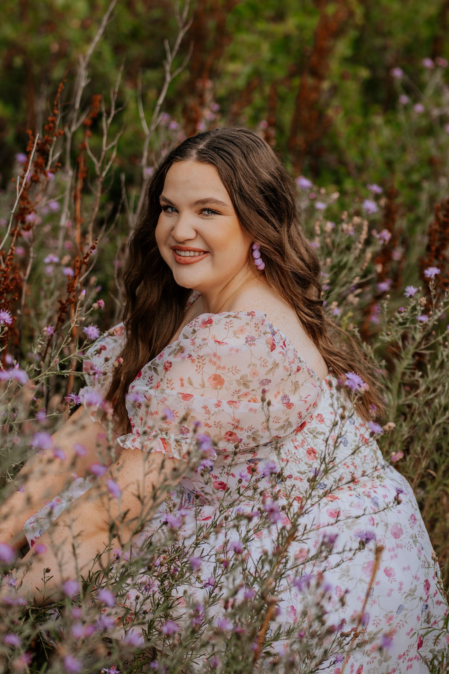 A smiling person in a floral dress poses among purple wildflowers in a natural outdoor setting during golden hour.