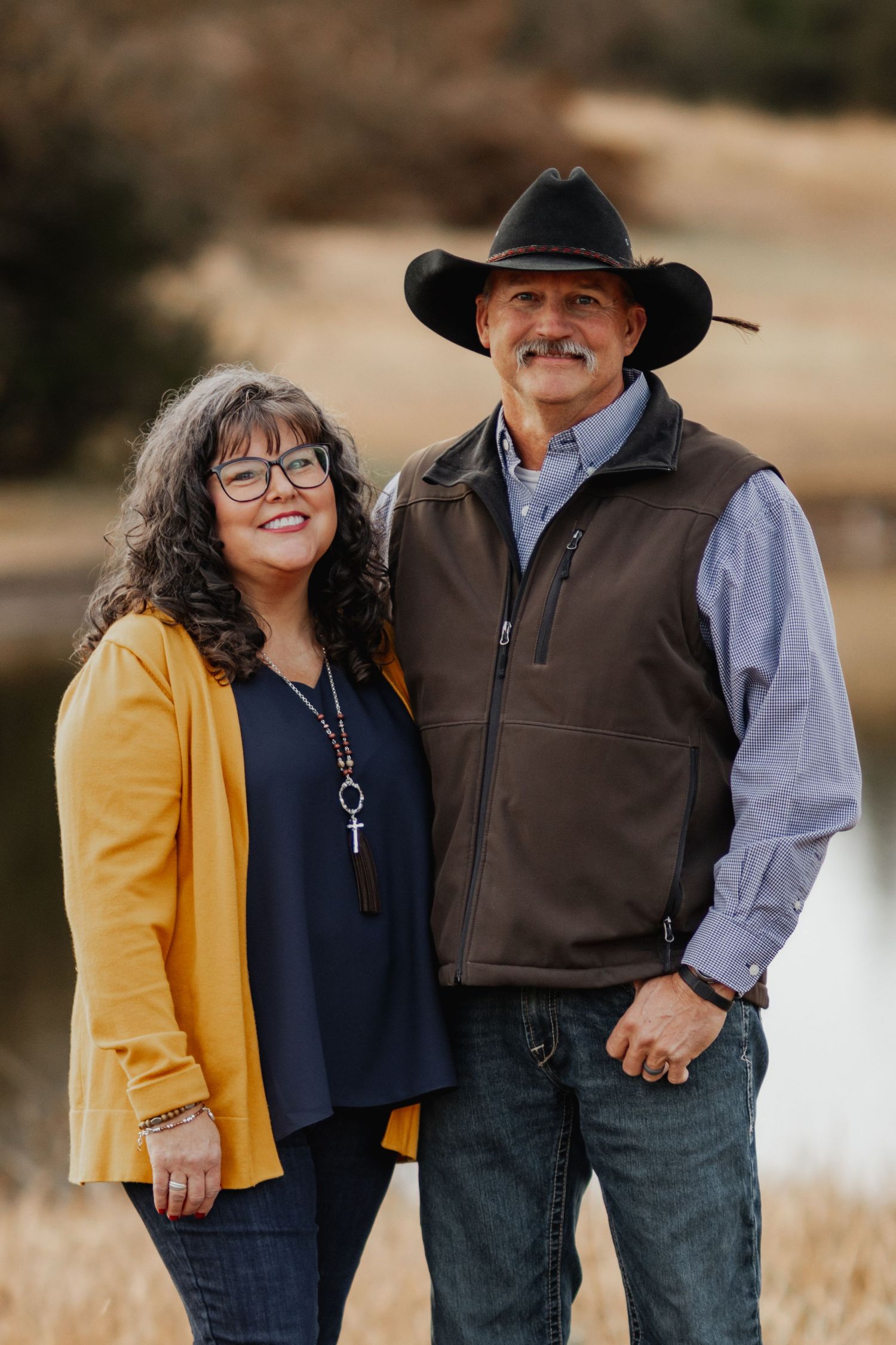 A couple stands together in a field wearing western attire including a cowboy hat and yellow cardigan.