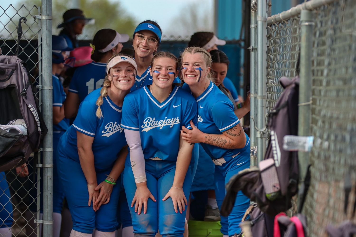 Softball team members share laughs in dugout wearing blue jerseys.