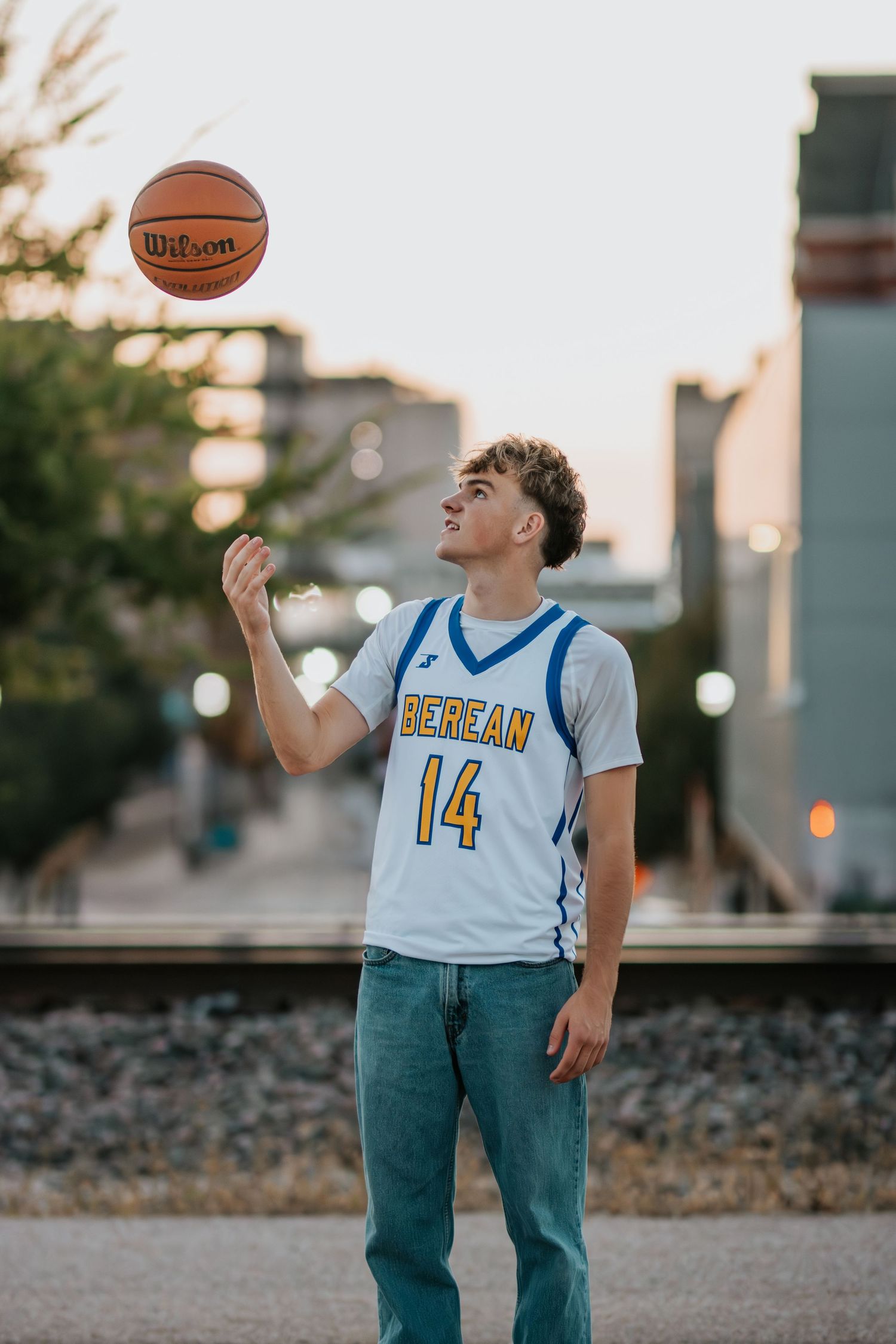 Basketball player in white jersey number 14 tosses ball in air on urban street at sunset.