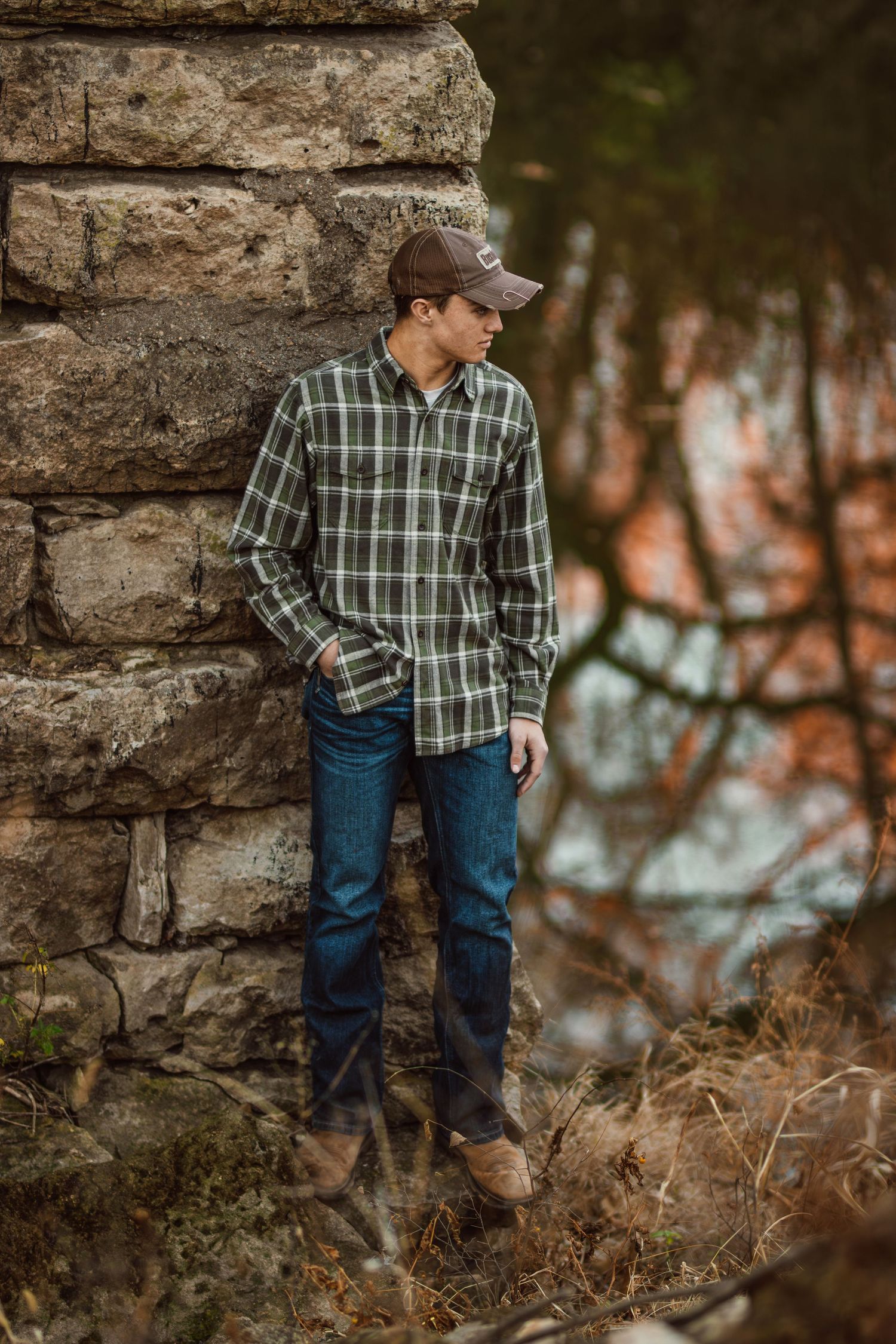 Person in plaid shirt and jeans leans against rustic stone wall in autumn setting.