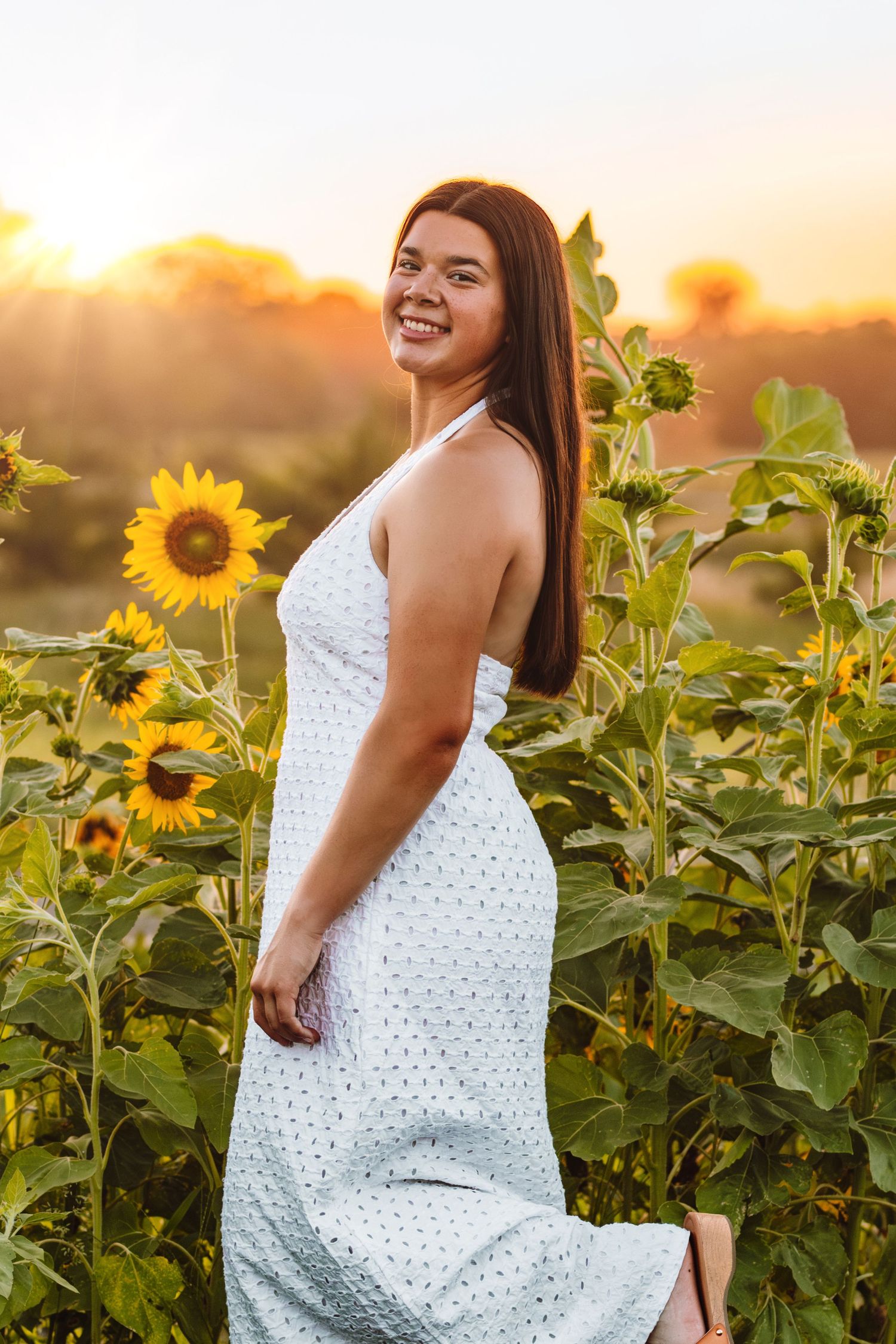 A joyful portrait in a white eyelet dress surrounded by tall blooming sunflowers at golden hour sunset.