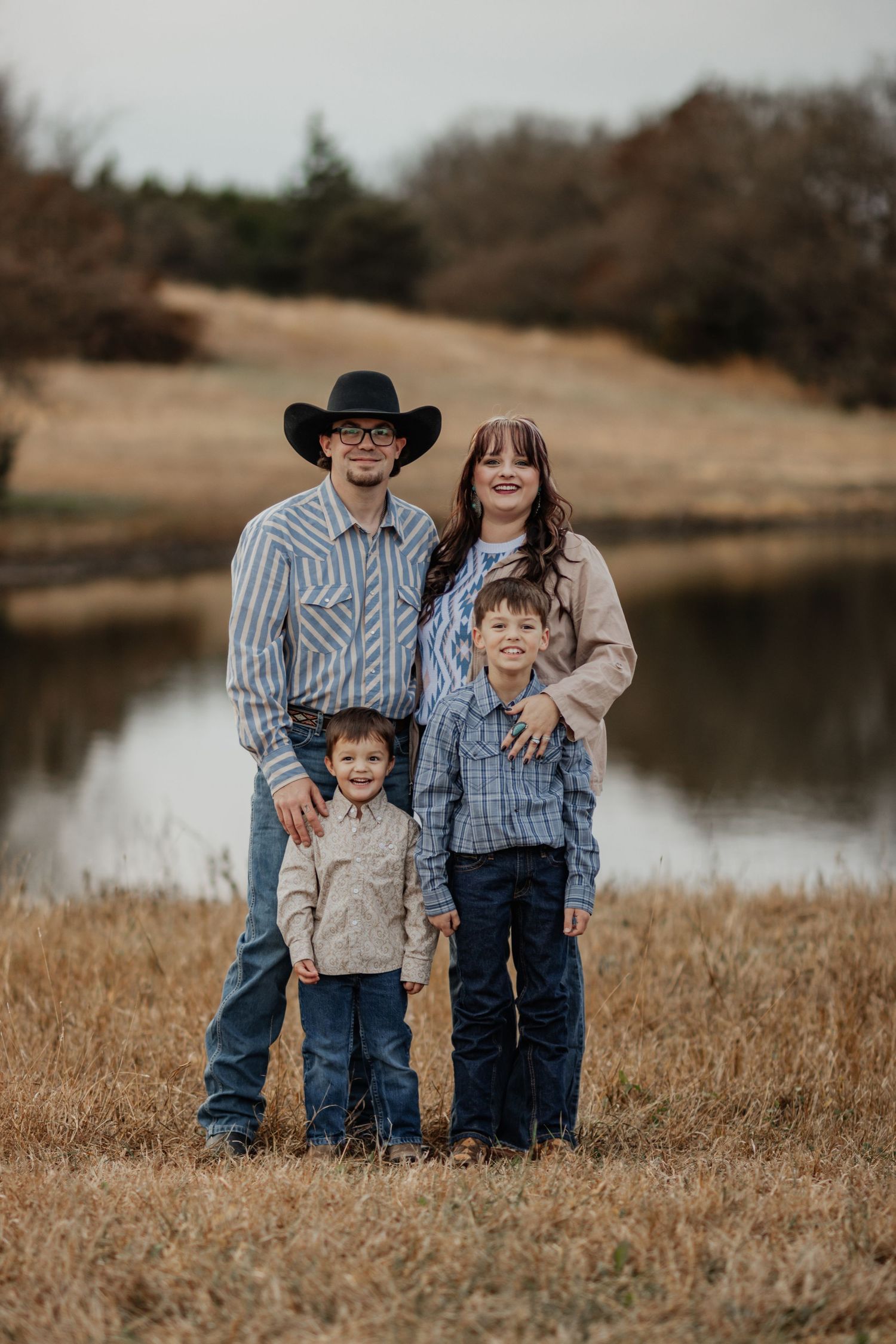 A family poses together in casual western wear by a pond on an autumn day with brown grass and trees in background.