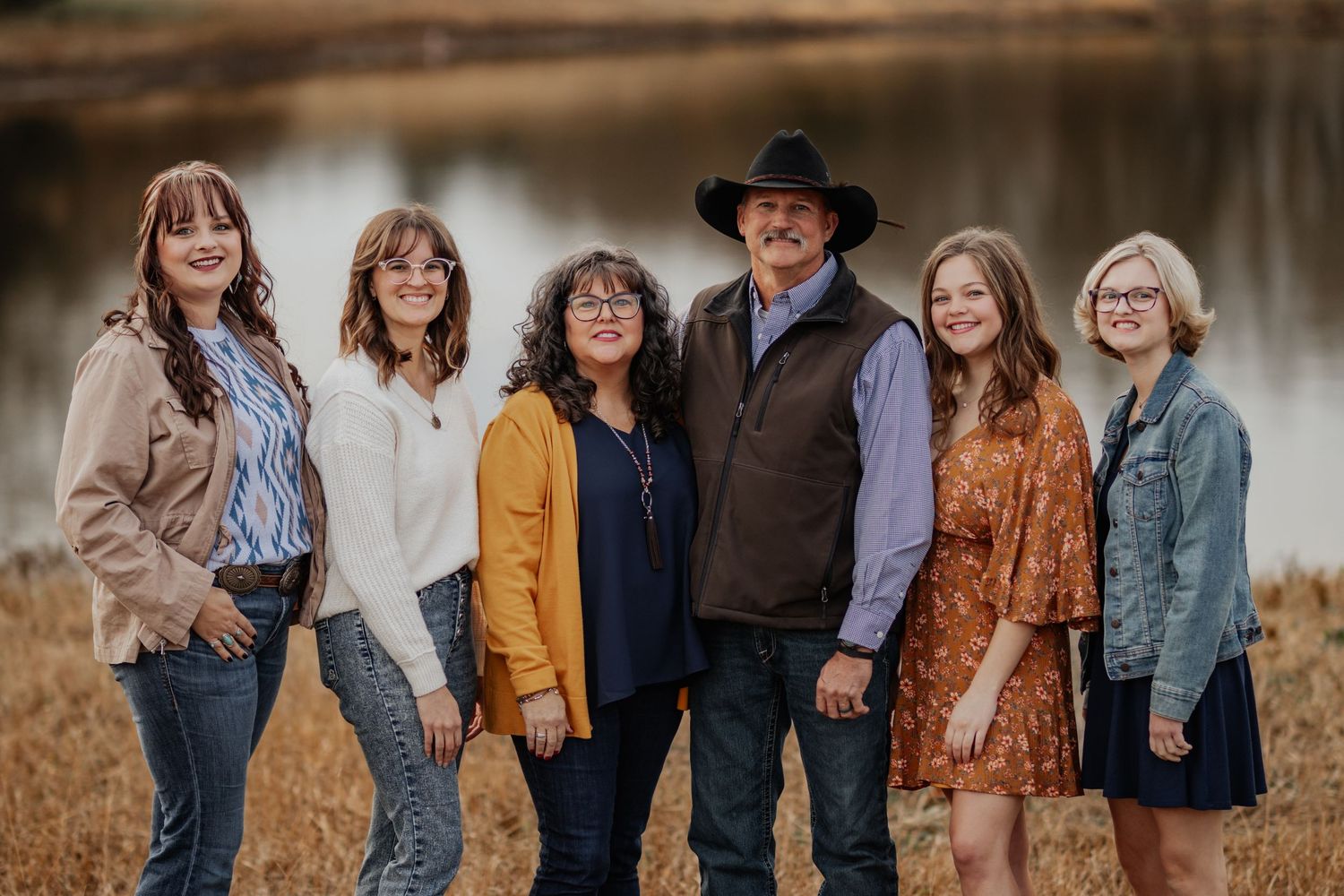 A group of six people wearing casual fall attire pose together outdoors by a serene lake at sunset.