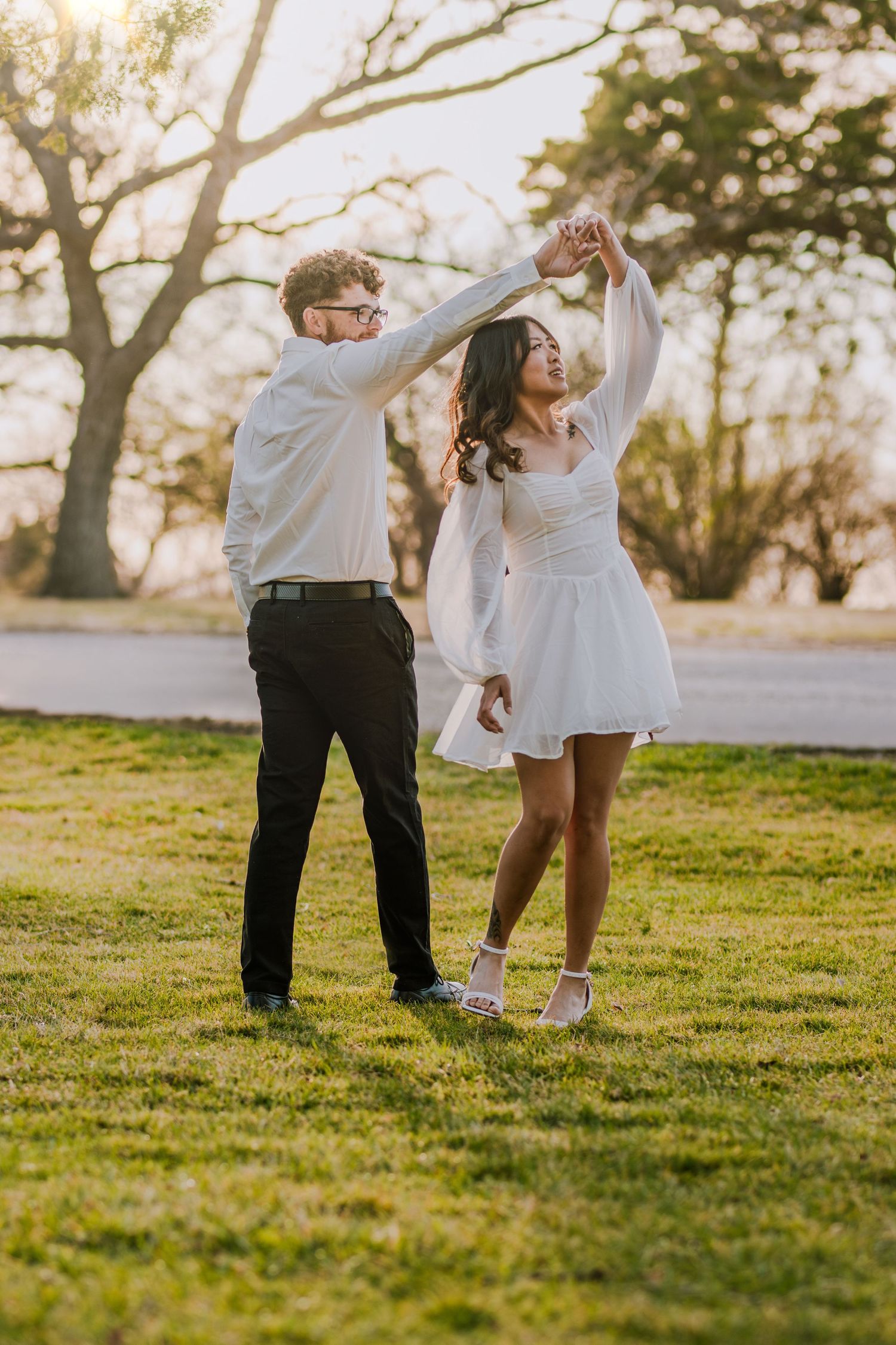 Couple dancing together in white outfits during sunset in park setting.