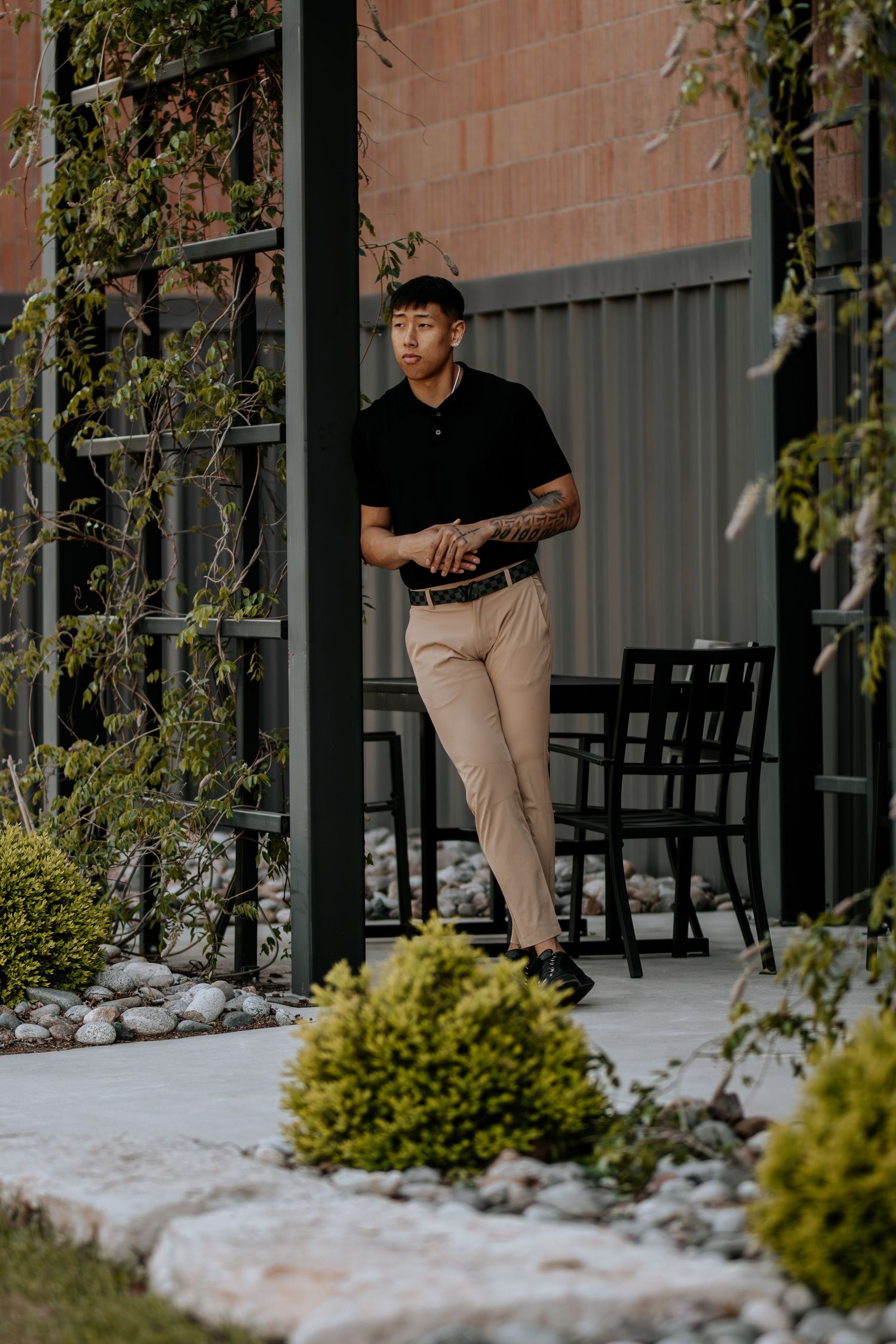 Person in black polo and khaki pants stands by landscaped building entrance.