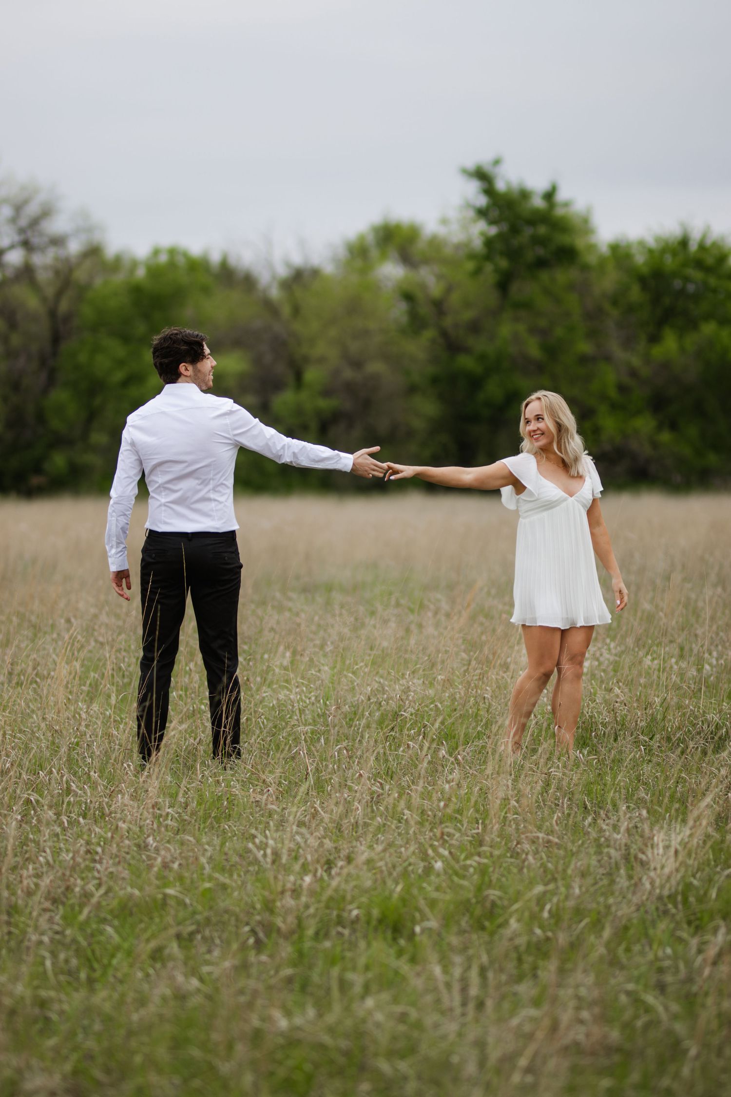 Couple in formal attire holds hands while standing apart in a rustic field with trees in the background.