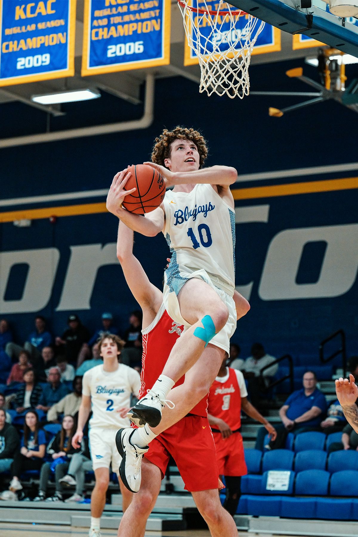 Basketball player in white Bluejays jersey leaps for layup against red-uniformed opponent.