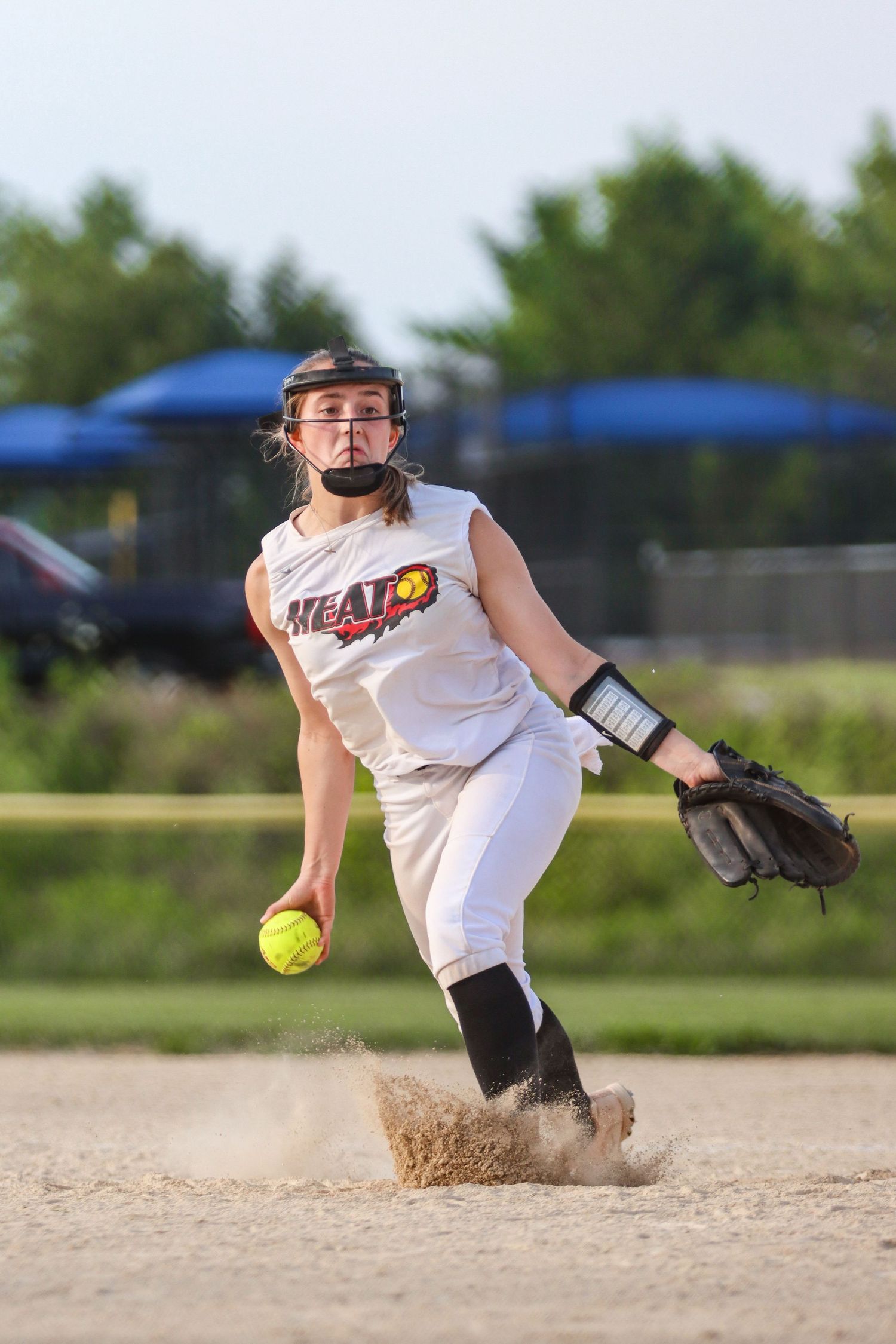 Softball pitcher in white uniform releases pitch while kicking up dirt on field.